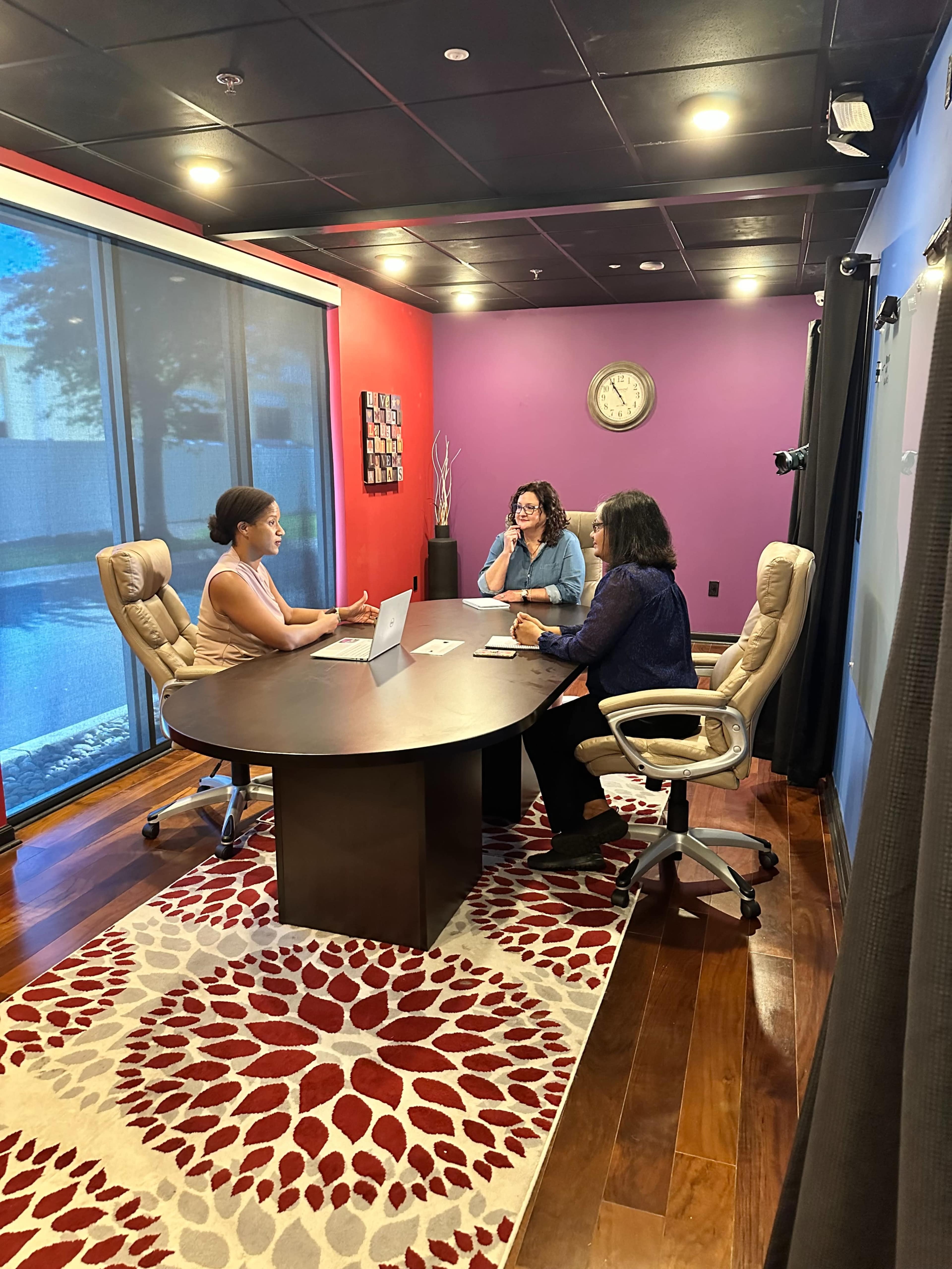 Three people sit around a large round table in a conference room, engaged in a discussion, with a laptop open in front of one of them and large windows allowing natural light.