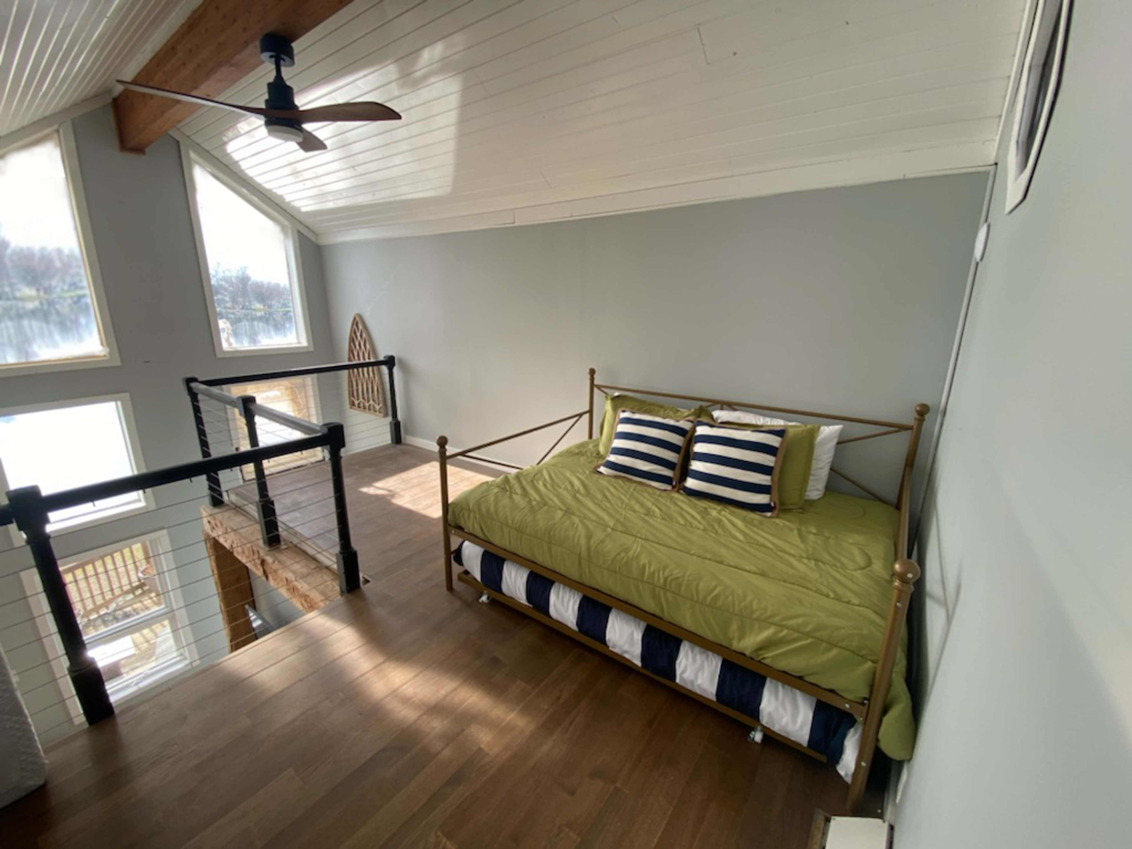 A cozy loft area features a green bedding set on a brass bed frame, large windows, and wooden flooring.