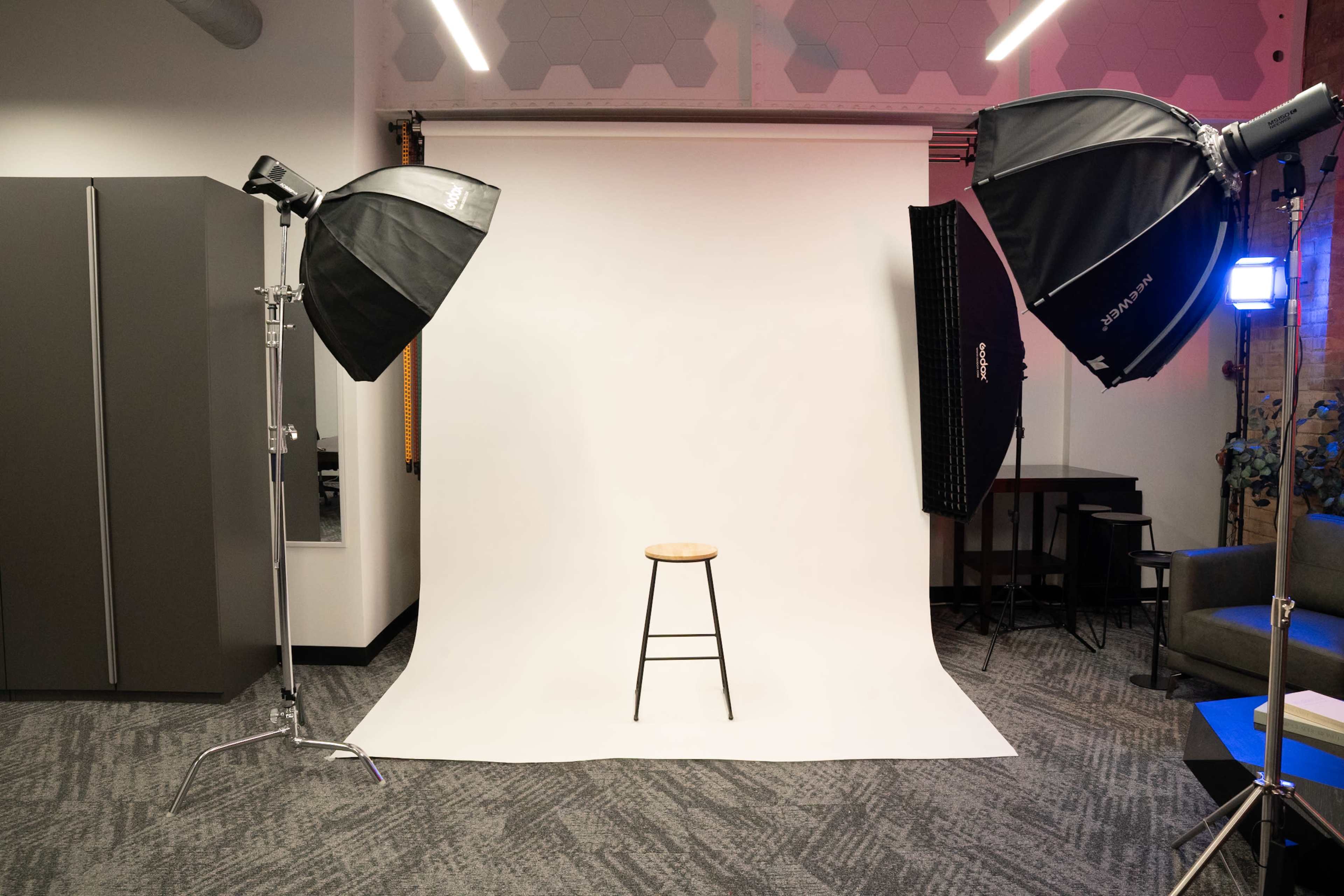 The image shows a photography studio setup featuring a plain white backdrop, a wooden stool in the center, and two large softbox lights positioned on either side.