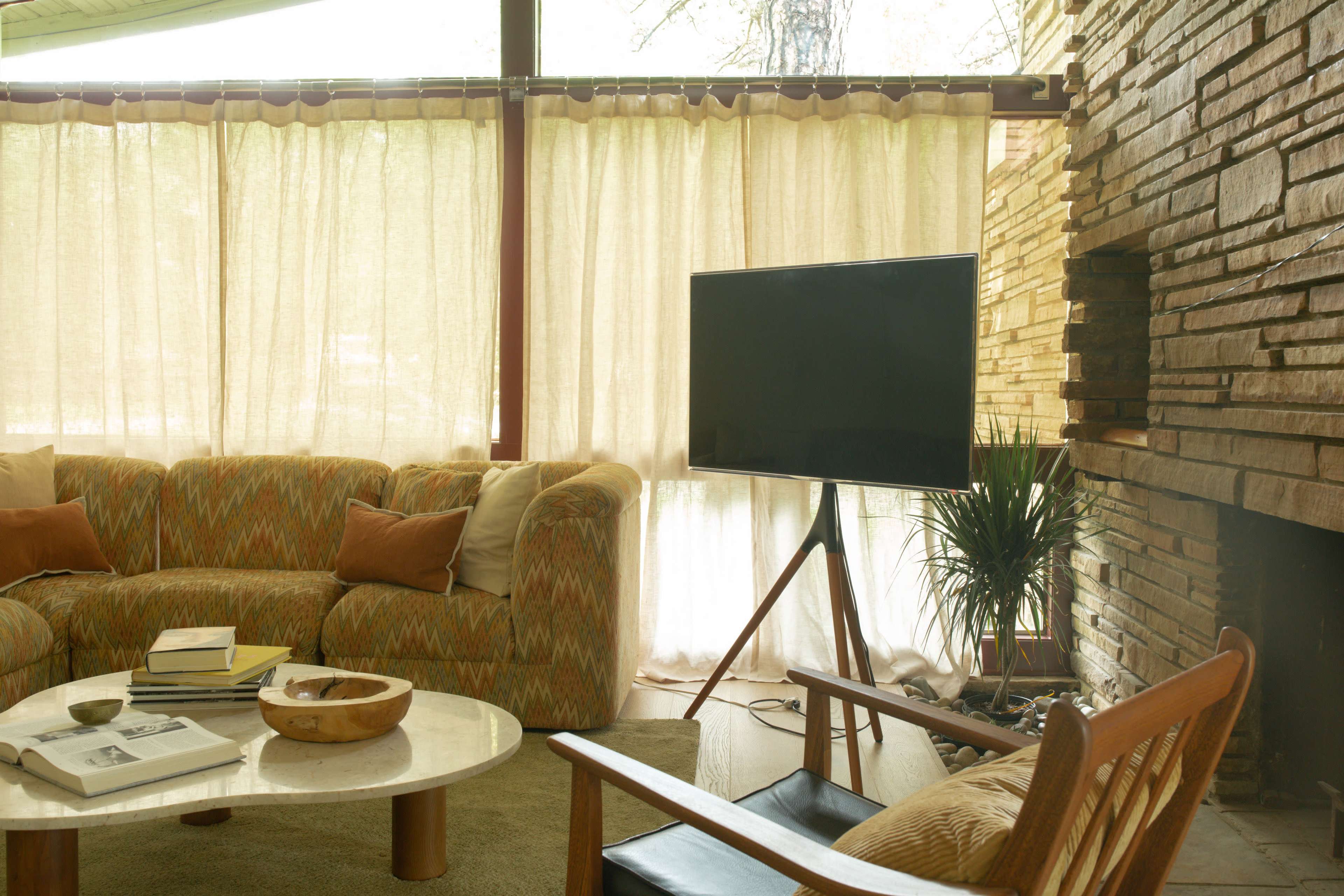 A cozy living room features a patterned sofa, a wooden chair, a flat-screen television, and a stone fireplace, all illuminated by natural light filtering through sheer curtains.