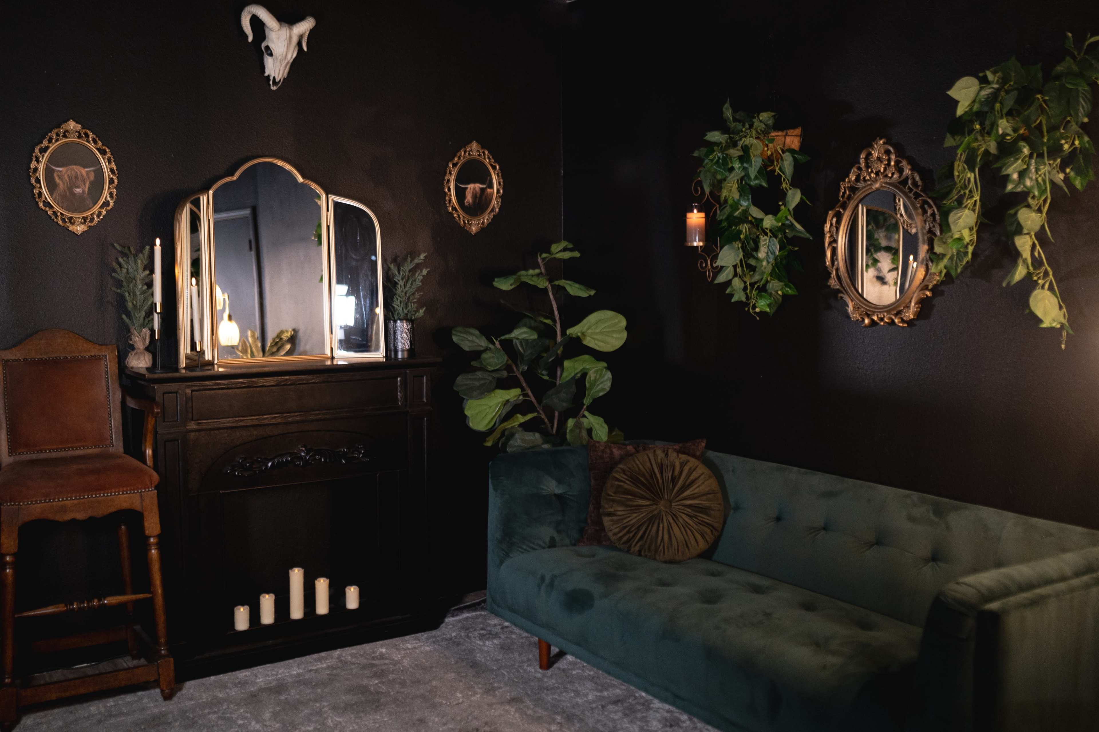 A dark room featuring a green velvet sofa, a wooden cabinet with candles, decorative mirrors, and wall decor including a skull and plants.