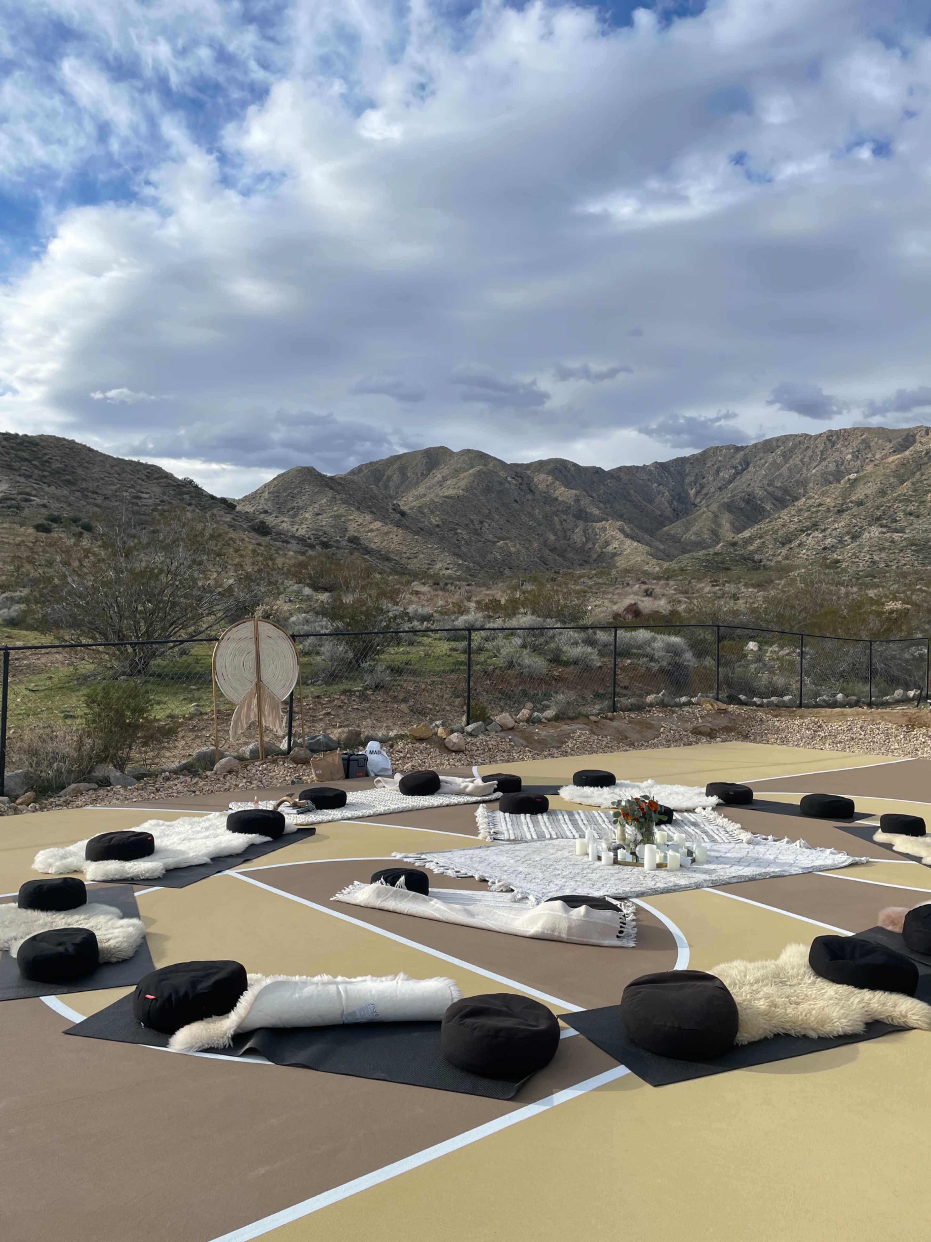 The scene depicts a meditation setup on a basketball court surrounded by mountains, featuring circular cushions arranged in a circular formation with soft textiles and candles in the center.
