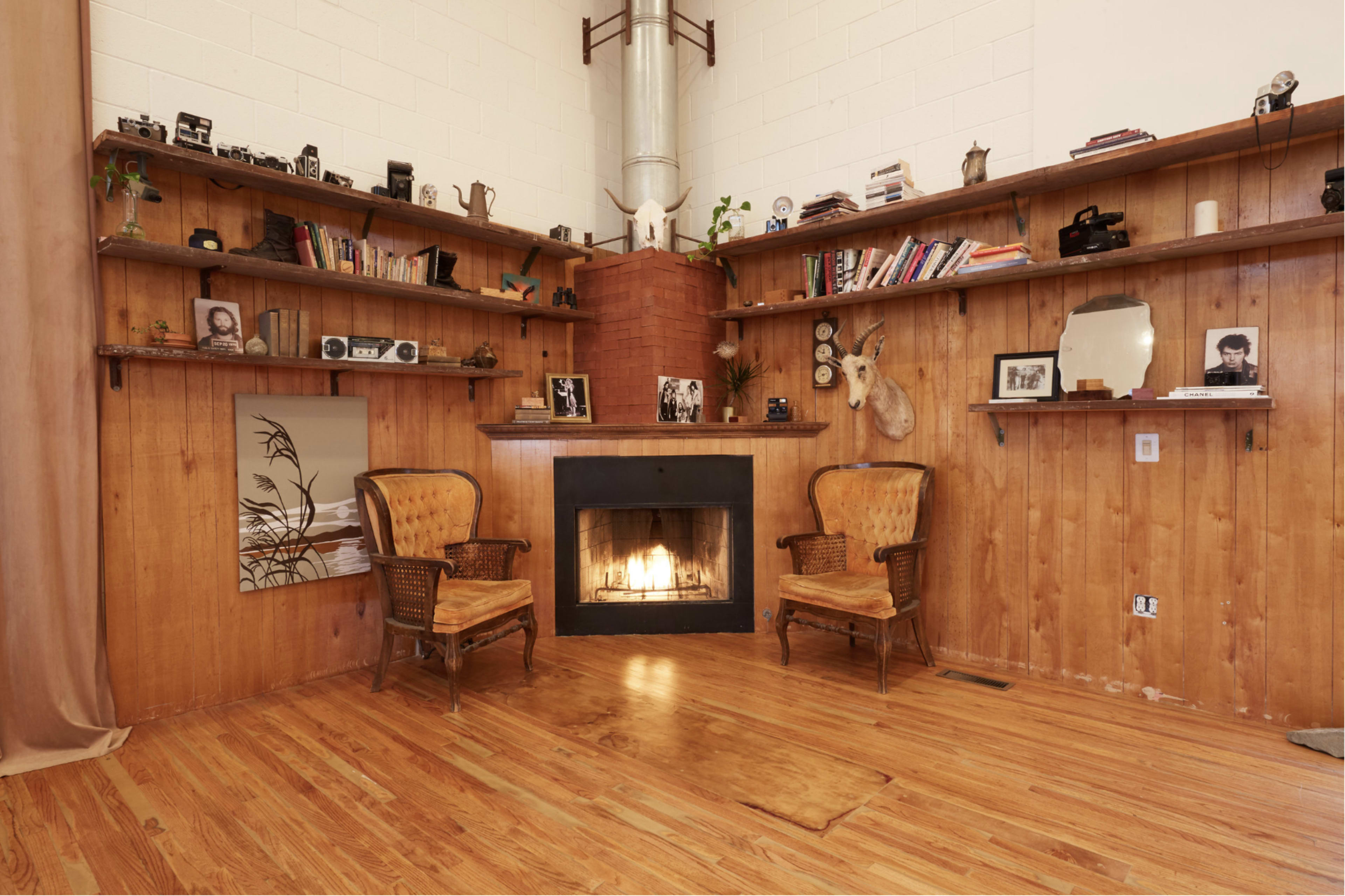 A cozy living room with two vintage chairs flanking a wood-burning fireplace, surrounded by wooden shelves filled with books and decorative items.