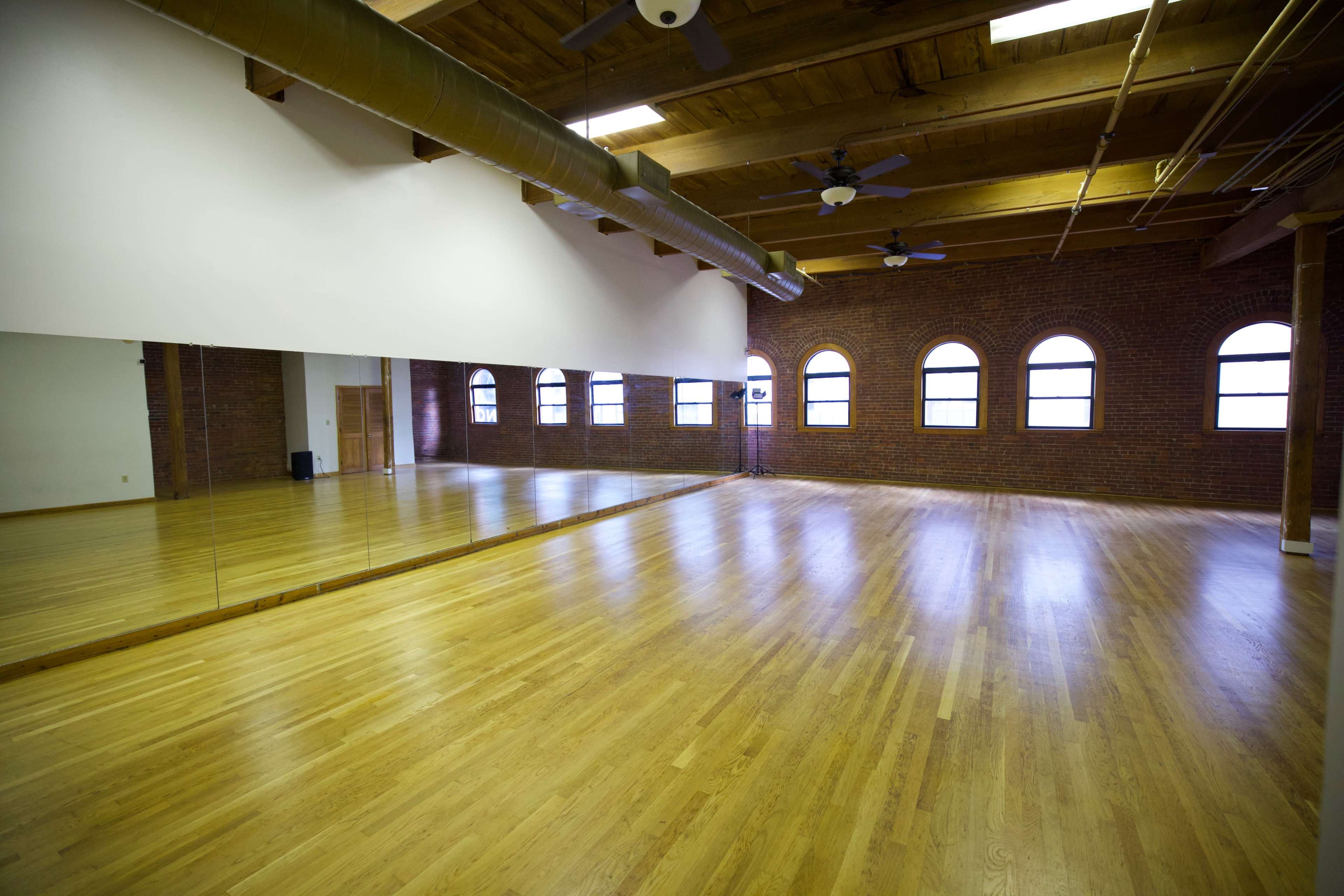 The image shows a spacious wooden-floored room with large windows and a wall of mirrors.