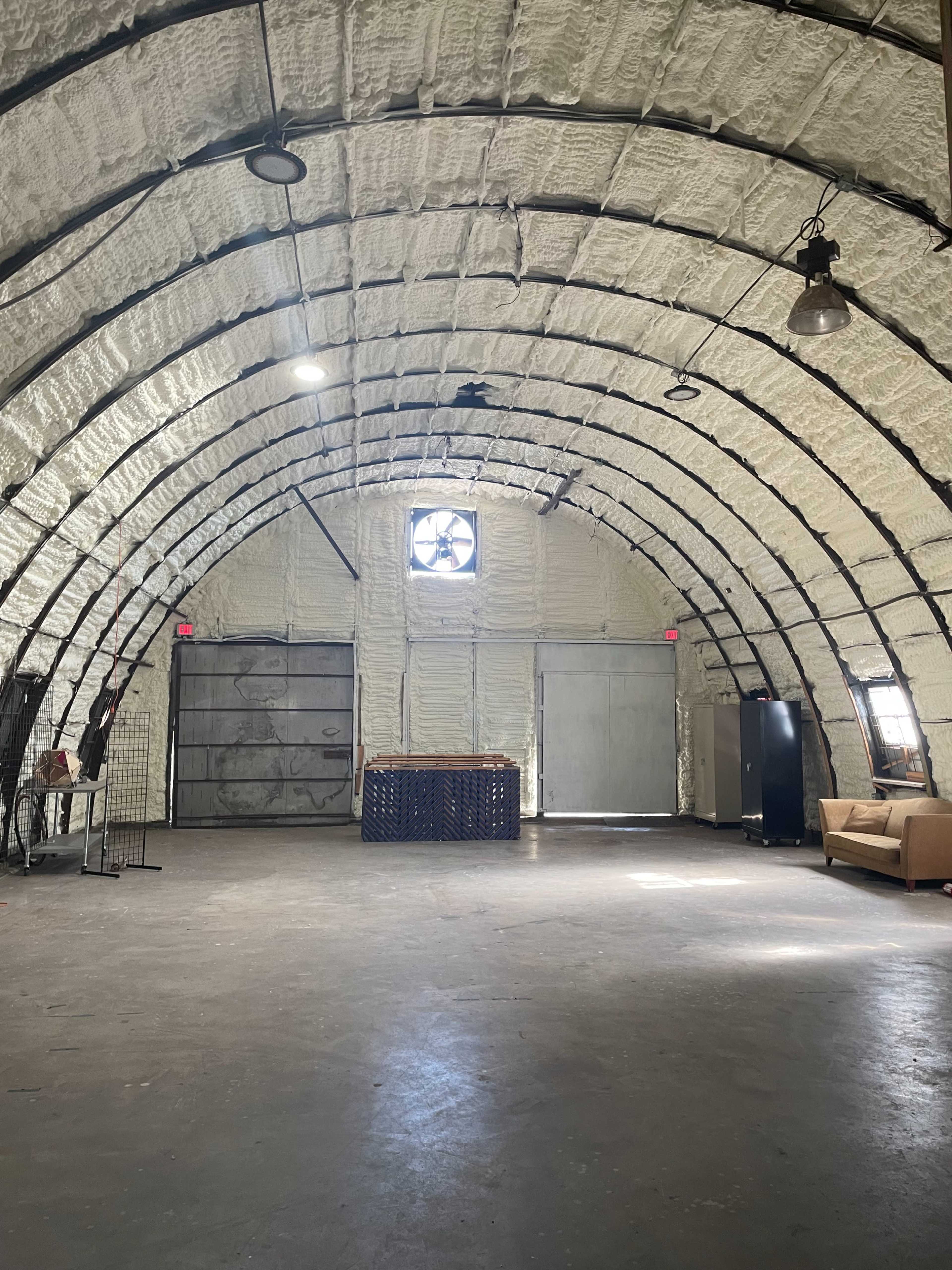 The interior of a large, arched building features exposed insulation, a fan in the window, and a couch against one wall.