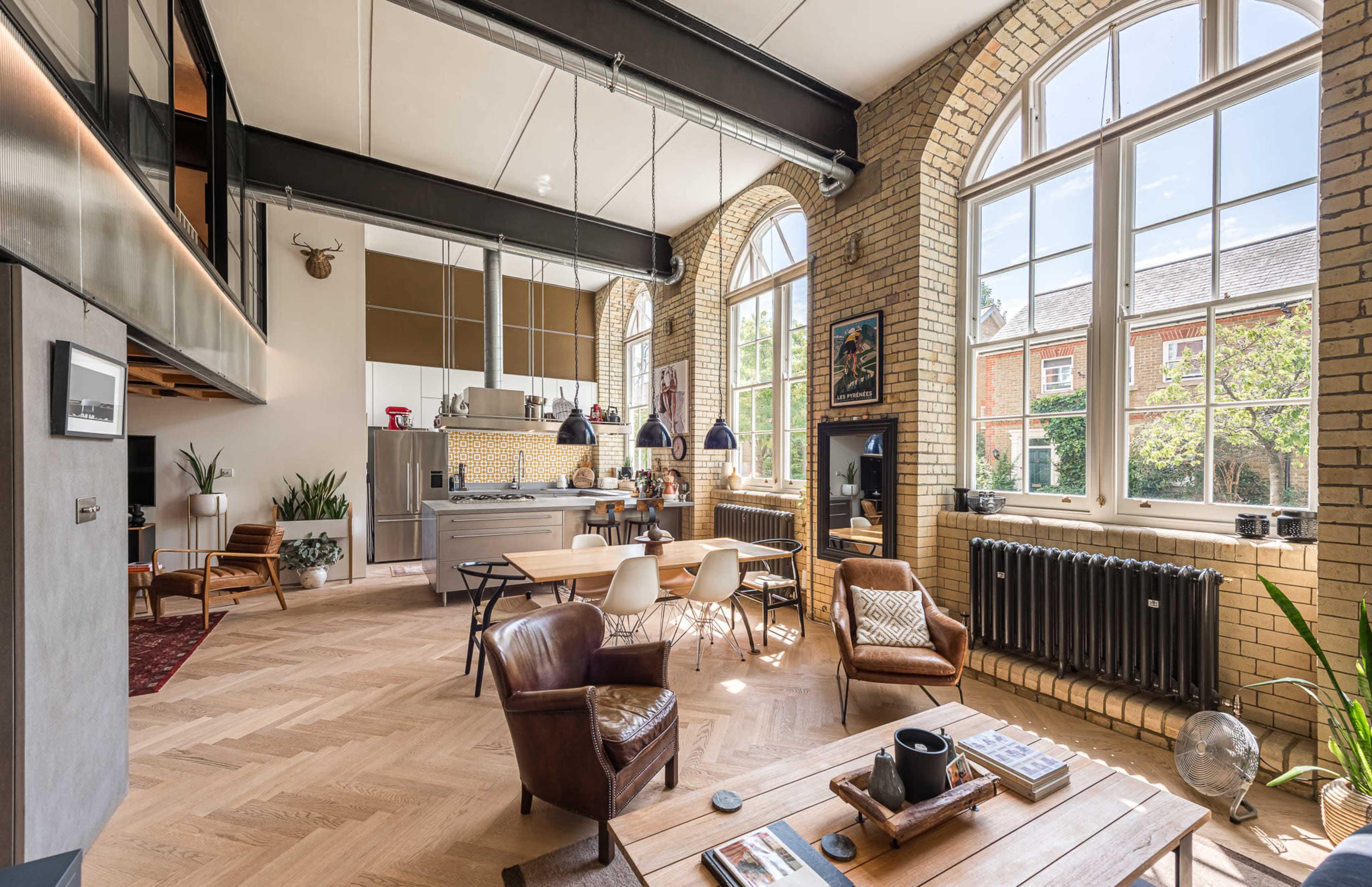 The image shows an open-concept living space featuring large windows, a modern kitchen area, a dining table, and a cozy seating arrangement with wooden flooring.