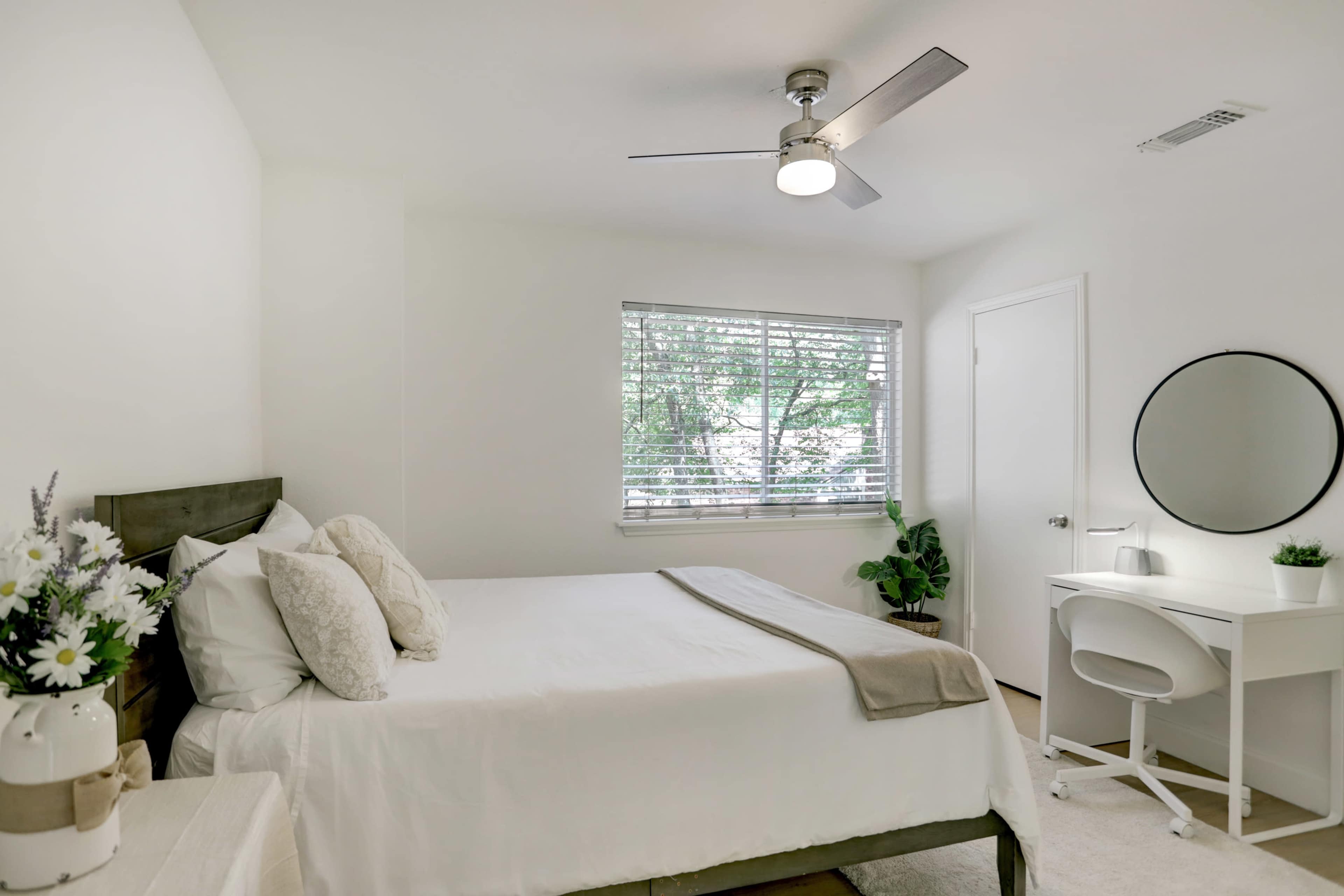 A simple bedroom features a bed with white linens, a nightstand with flowers, a desk with a mirror, and a window overlooking greenery.