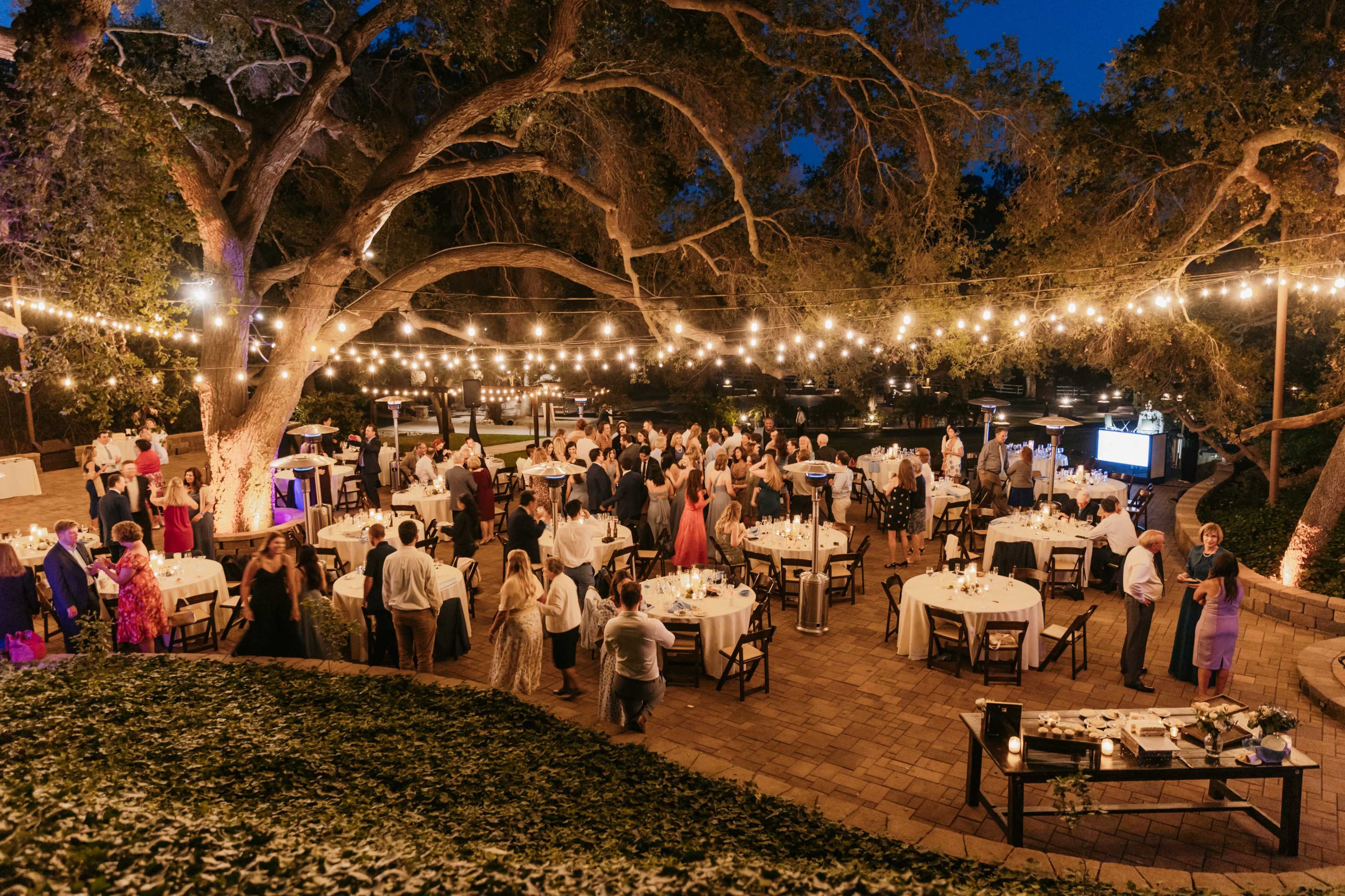 The image shows a large outdoor gathering at night, with string lights illuminating tables and guests mingling around a landscaped area.