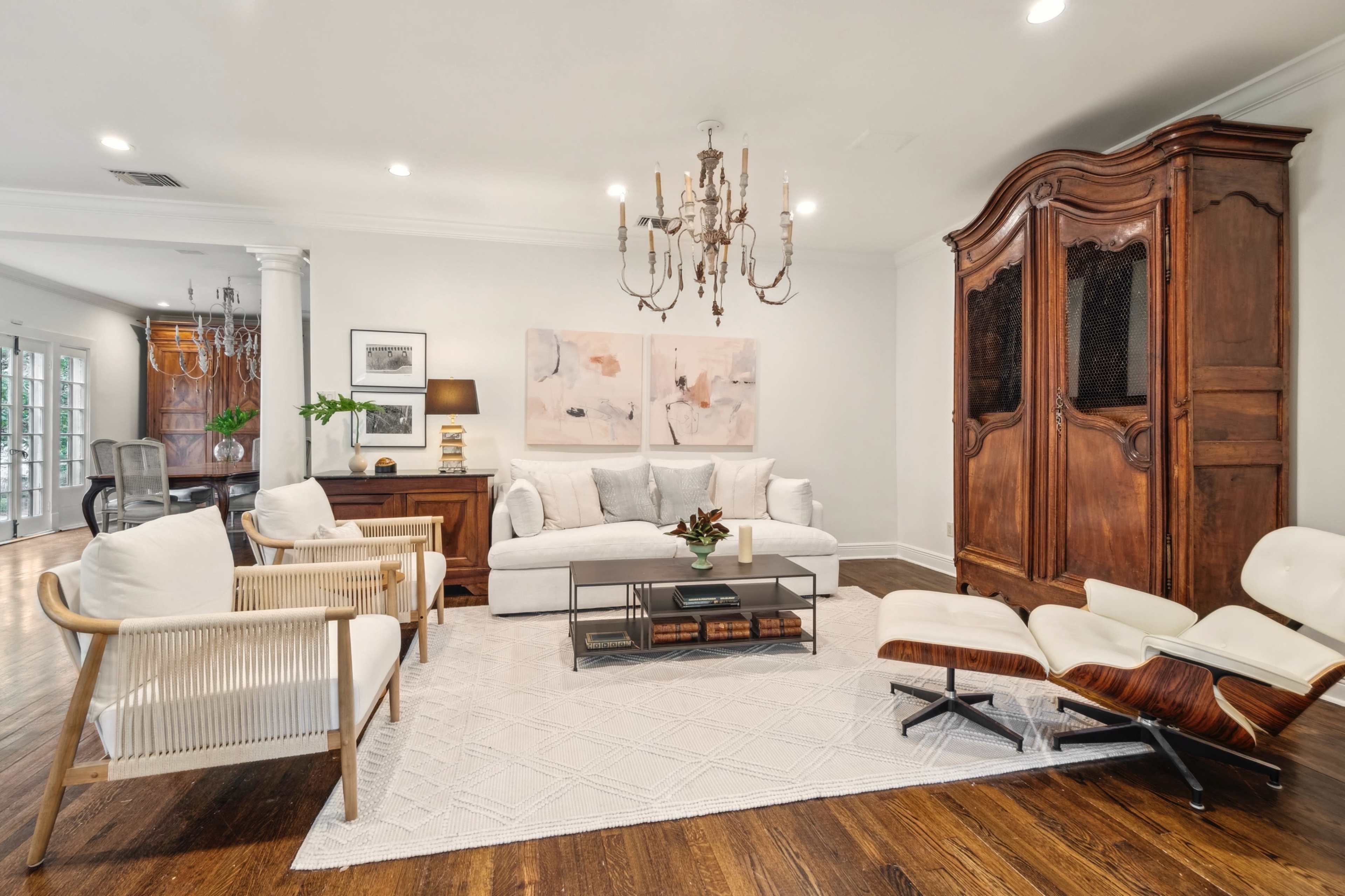 The image depicts a modern living room featuring a white sofa, two light-colored armchairs, a stylish coffee table, and a large wooden cabinet, all arranged on a textured rug with a chandelier overhead.