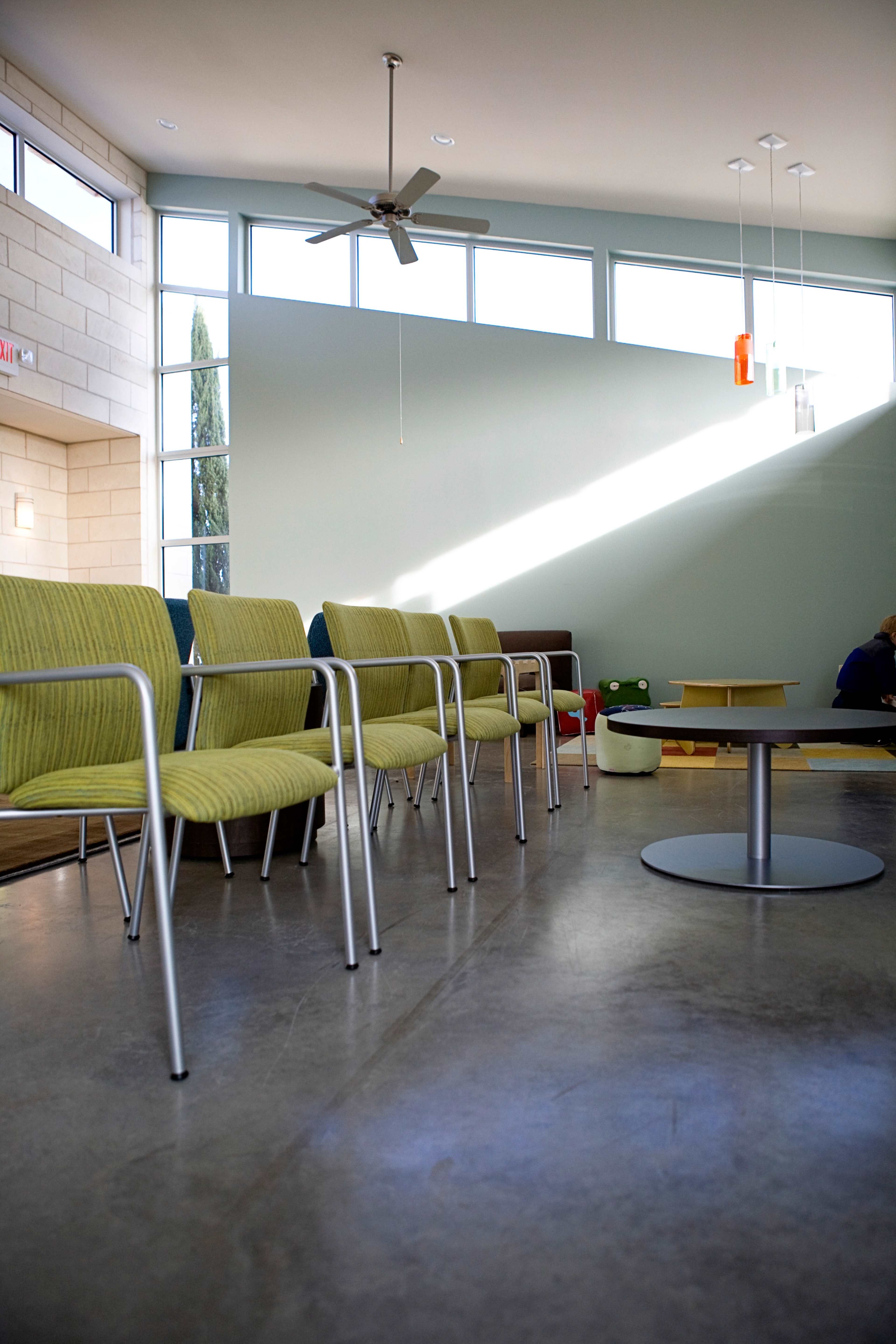 A modern waiting area with several green chairs arranged around a central table, illuminated by natural light from large windows.