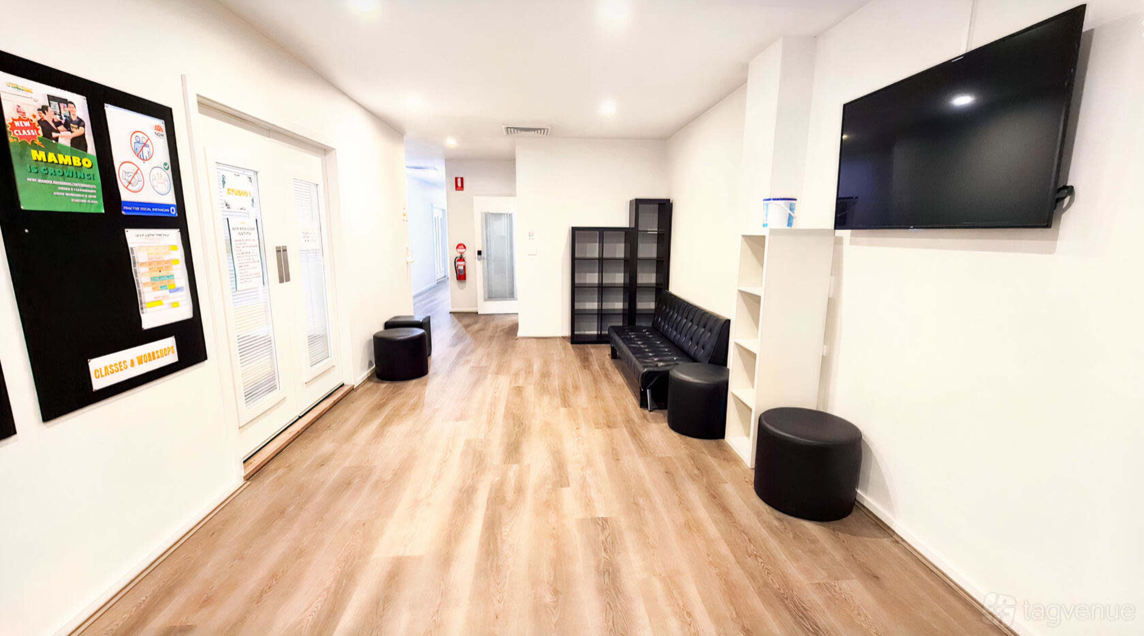 The image shows a modern, well-lit hallway with a black leather sofa, several round black stools, shelves, and a wall-mounted television.