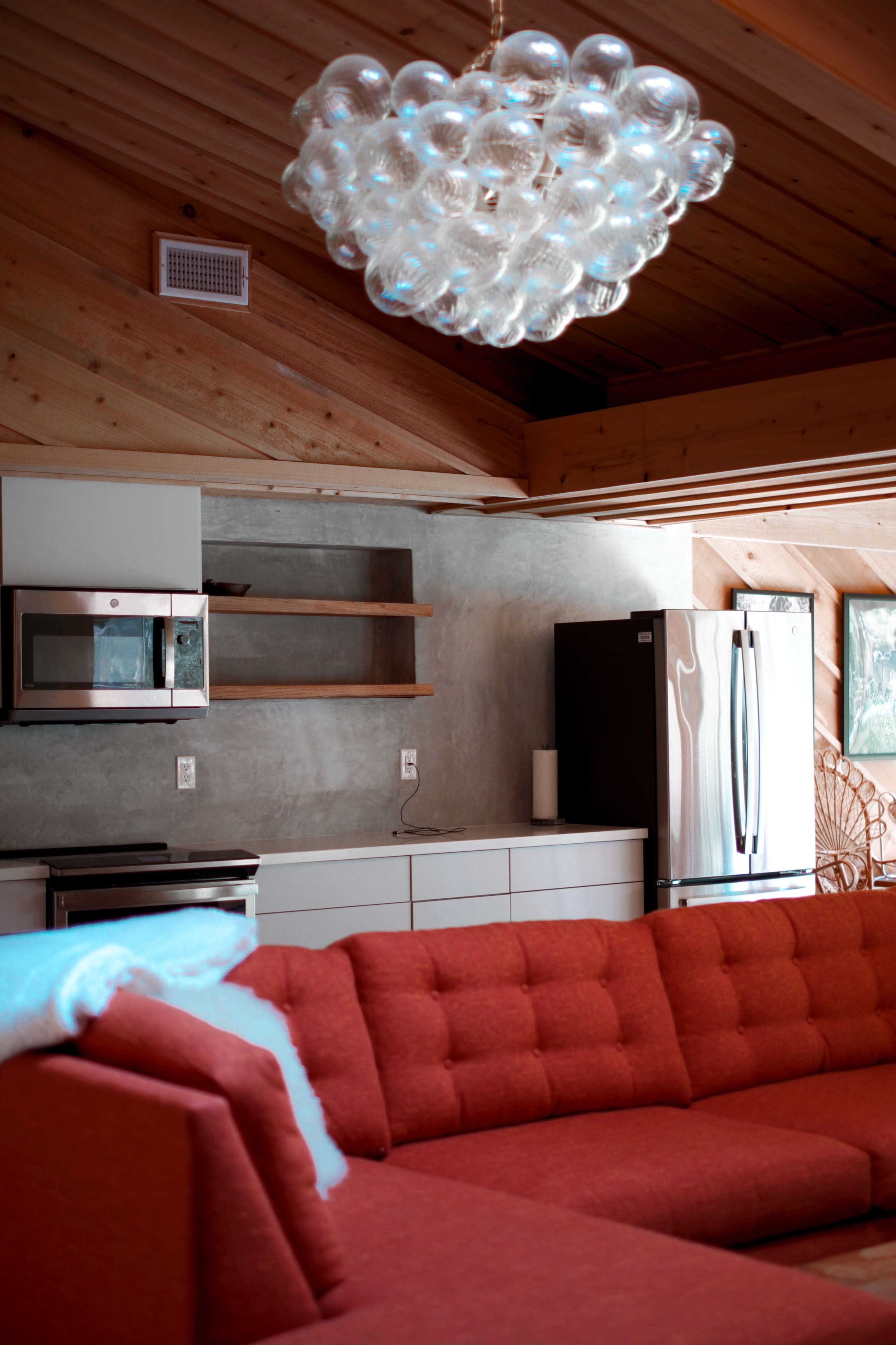 The image shows a modern living room featuring an orange sectional sofa, a bubble chandelier, and a kitchen area with a refrigerator and cabinetry.