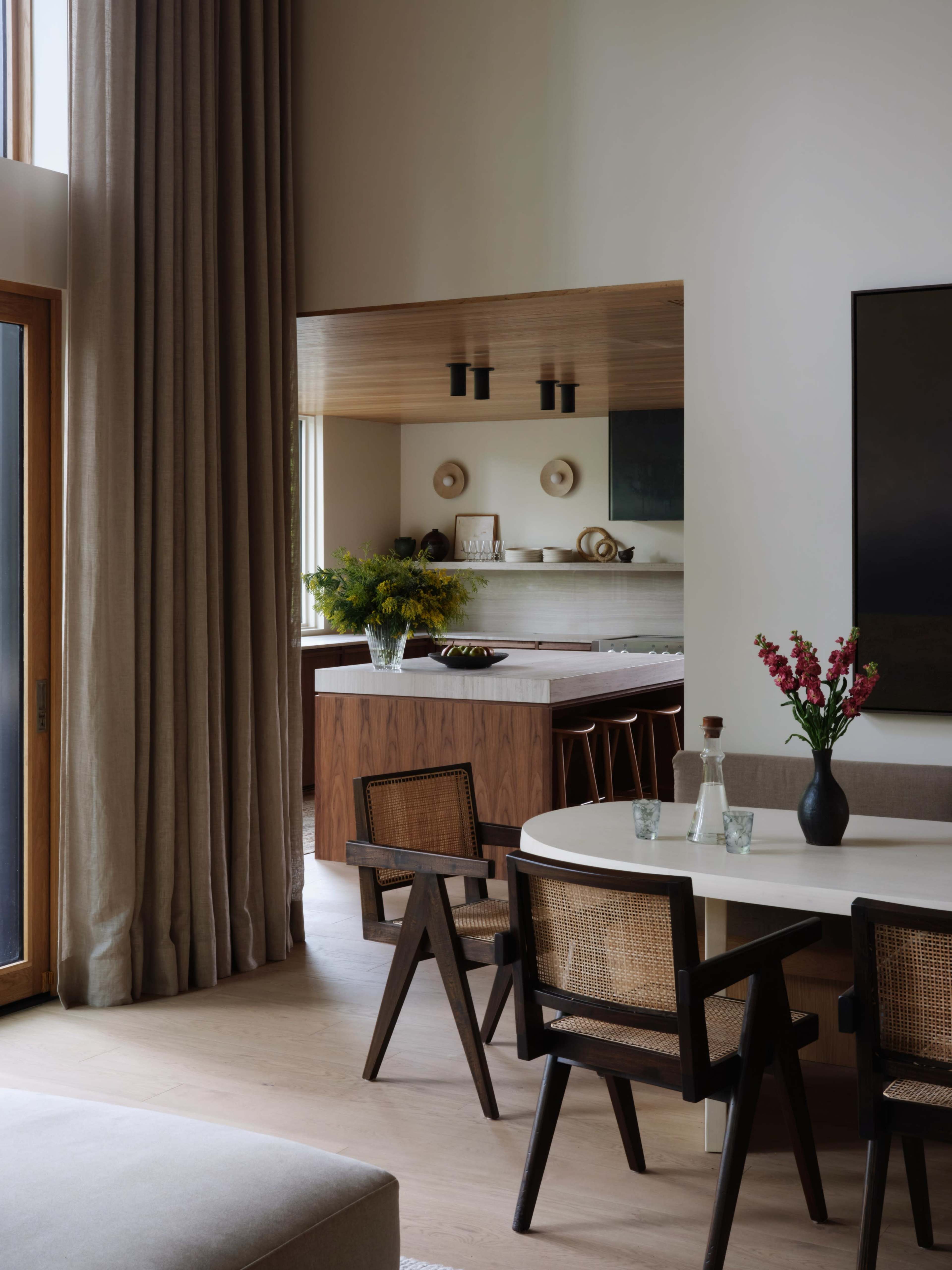 A modern interior features a dining area with a round table surrounded by wooden chairs, set against a backdrop of large windows and soft drapery.