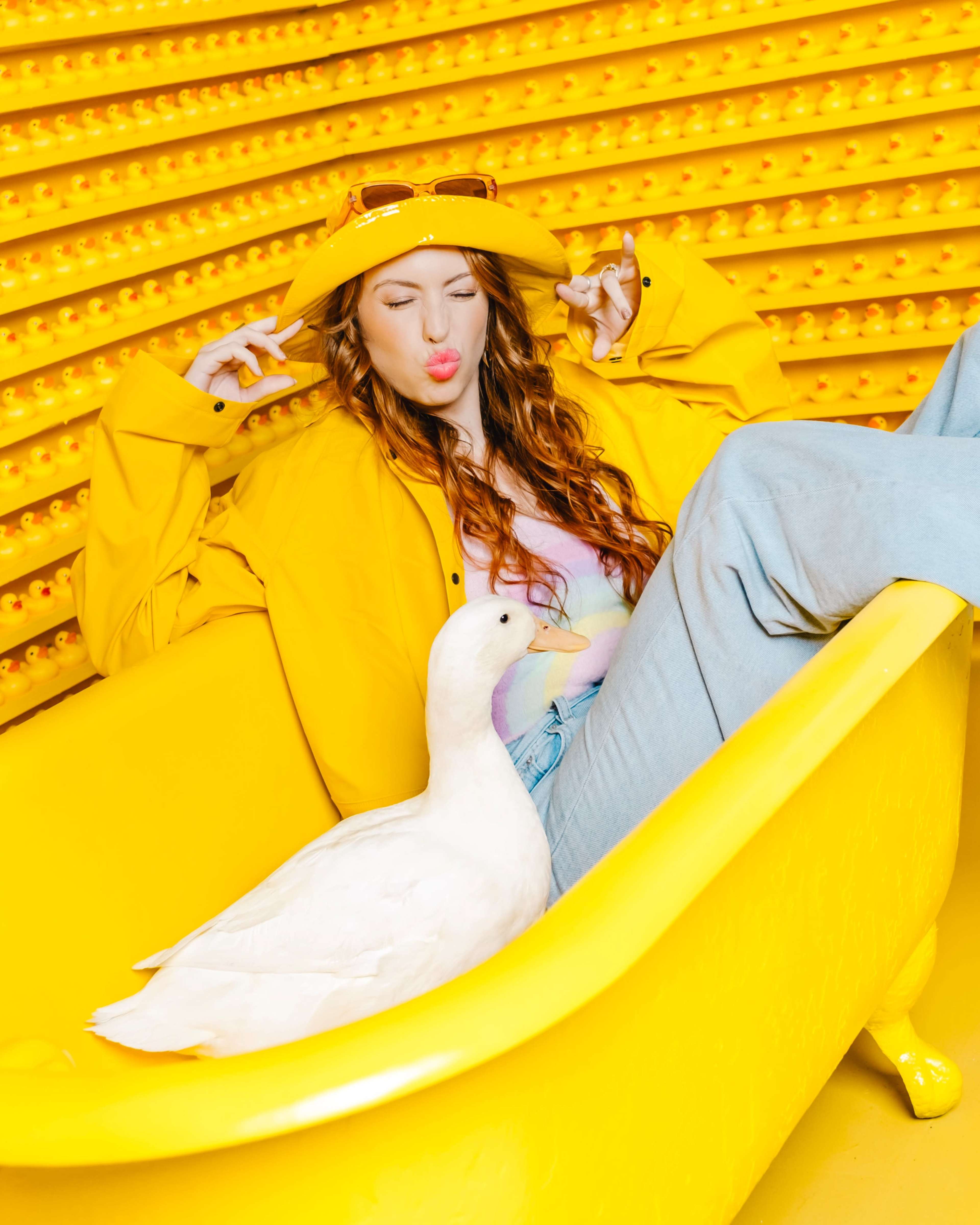 A person sits in a yellow bathtub wearing a yellow jacket and hat, playfully pouting while a duck is beside them.