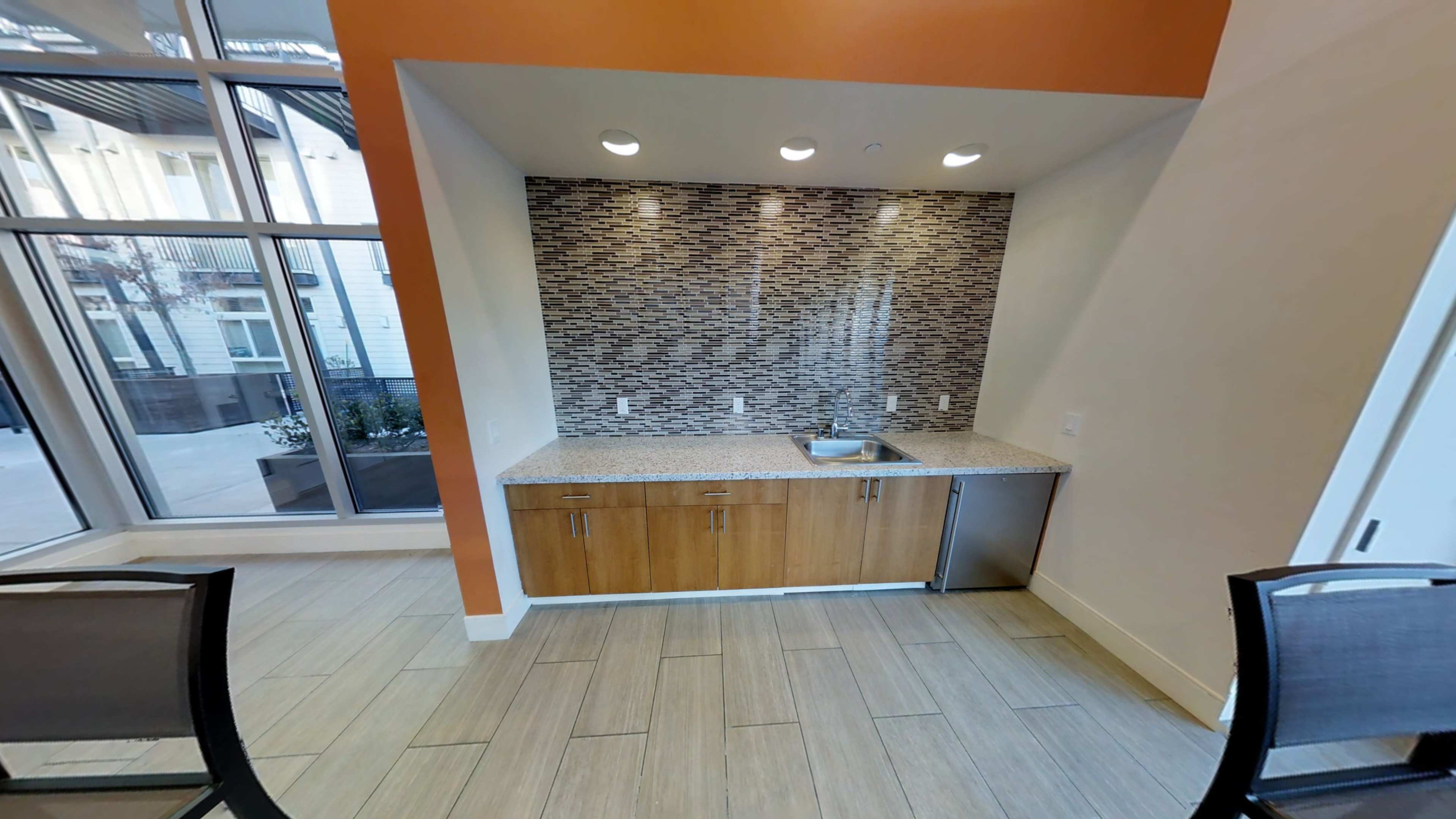 A small kitchenette area featuring a granite countertop, a sink, and a backsplash made of decorative tiles, situated in a room with large windows.