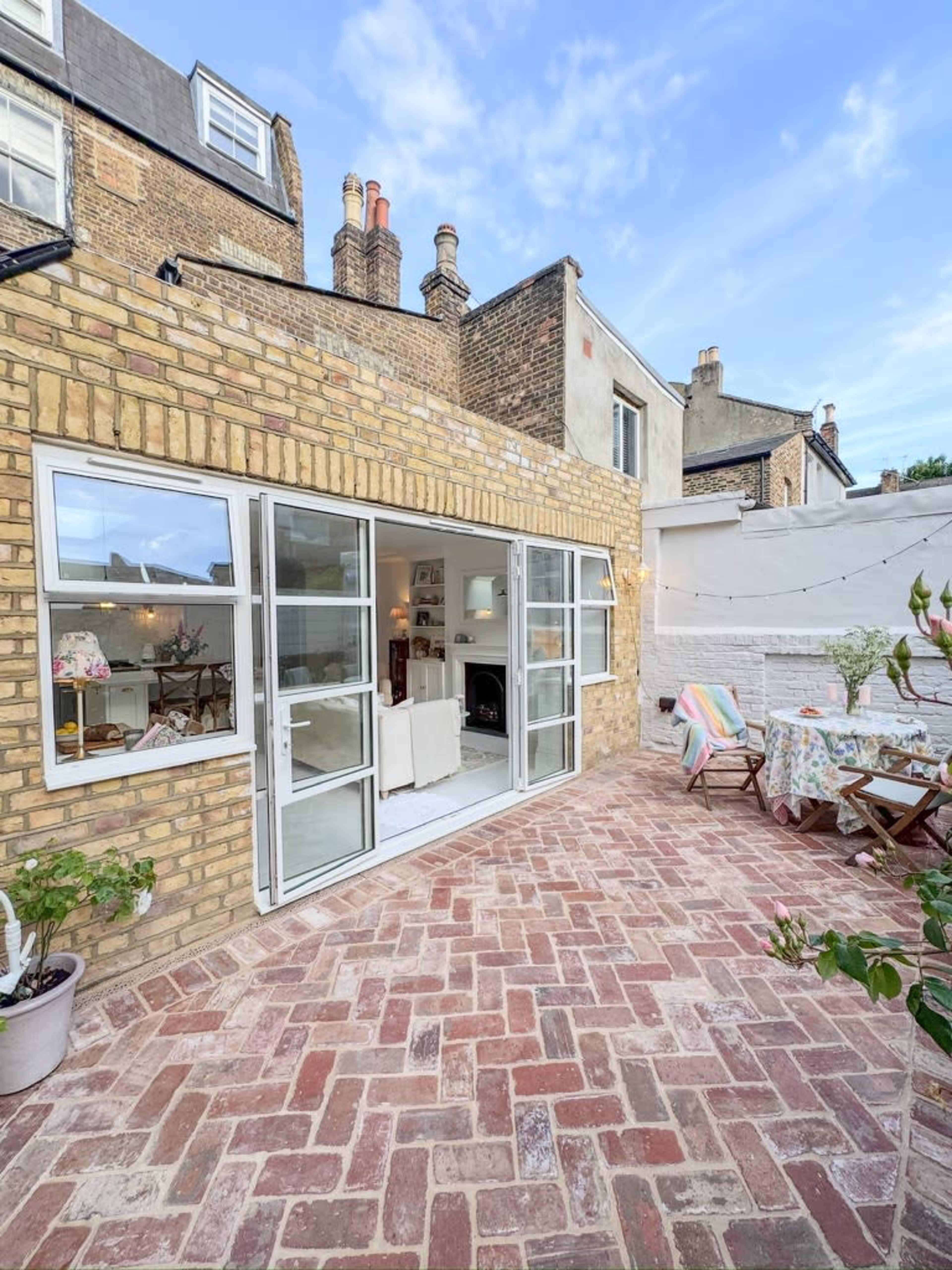 The image shows a brick-paved patio area that opens into a cozy interior space with large glass doors.