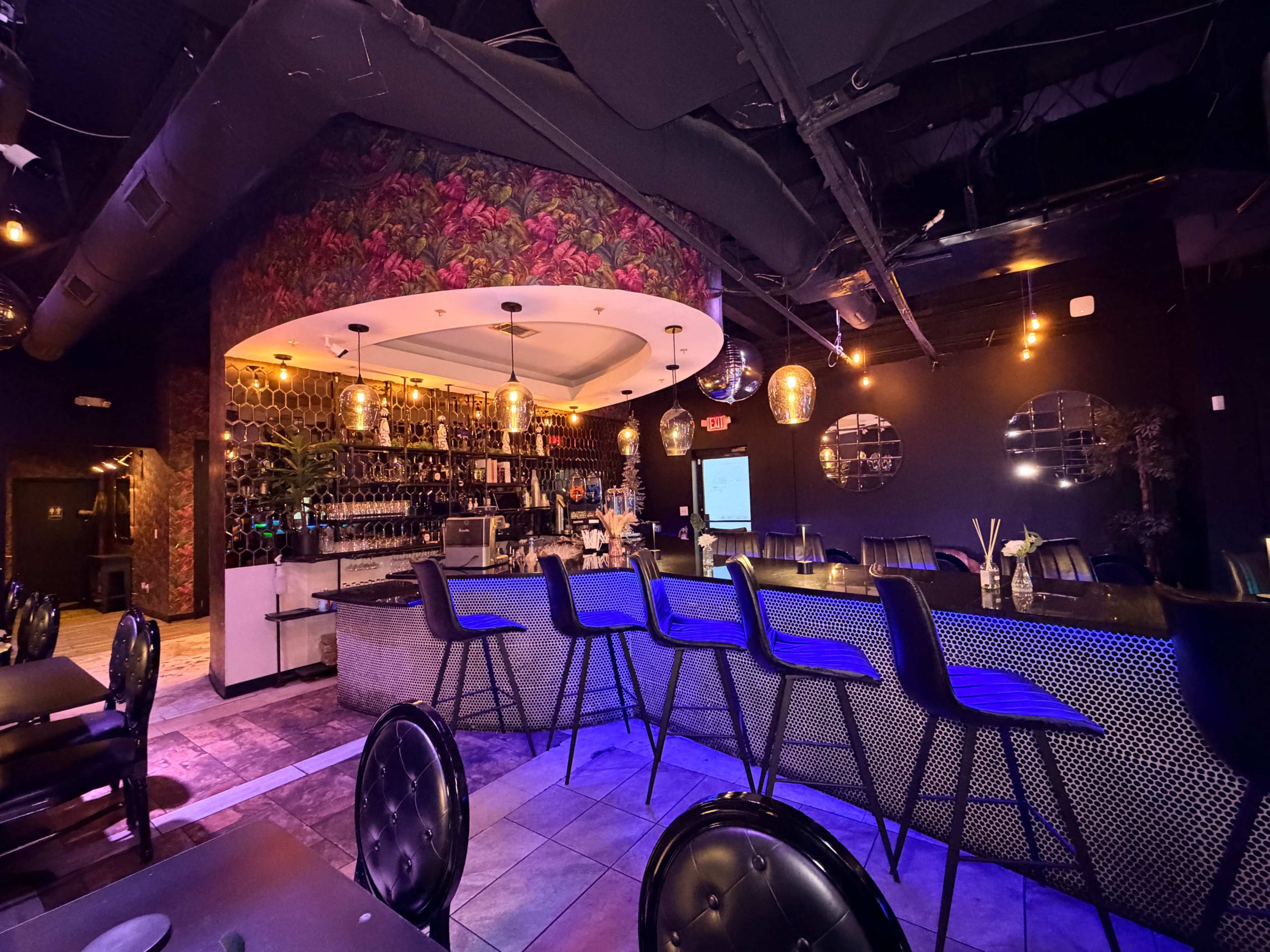 The image shows a stylish bar area inside a dimly lit restaurant, featuring a curved counter, high stools, and decorative floral wallpaper.