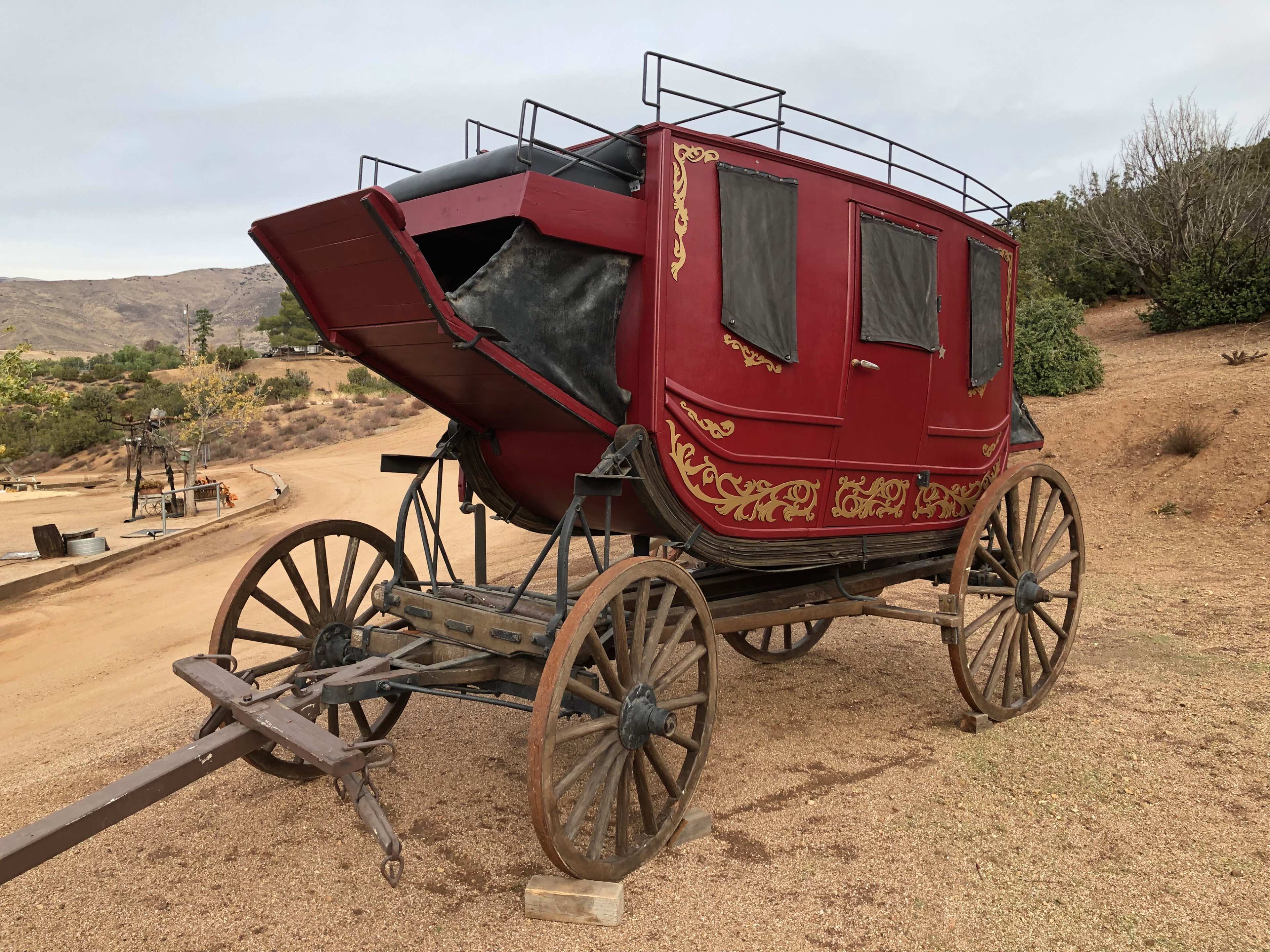 A vintage red stagecoach with decorative gold detailing, mounted on wooden wheels, positioned on a dirt pathway in a rural setting.