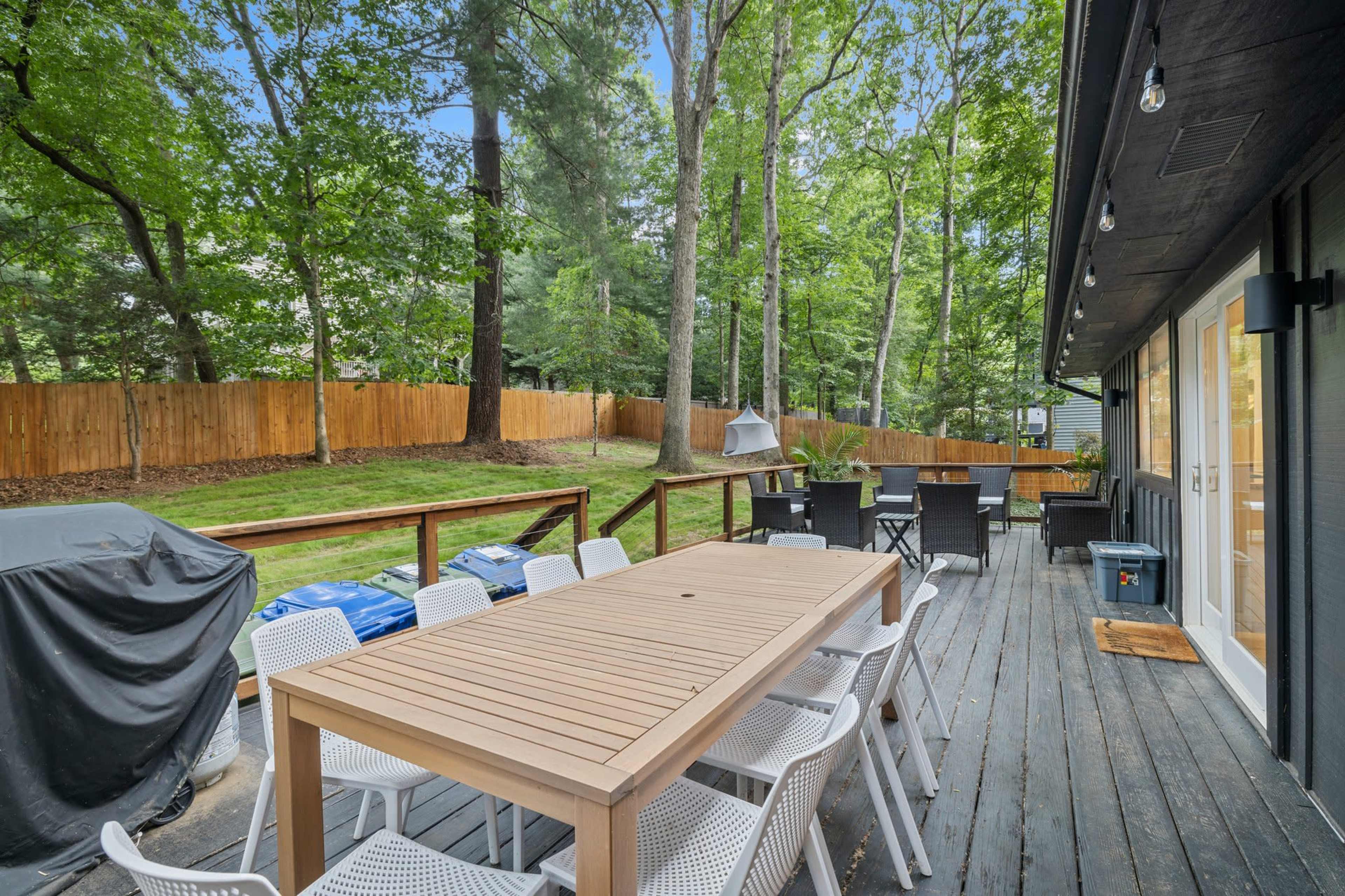 A wooden deck features a large table and white chairs, surrounded by trees and a fenced backyard.