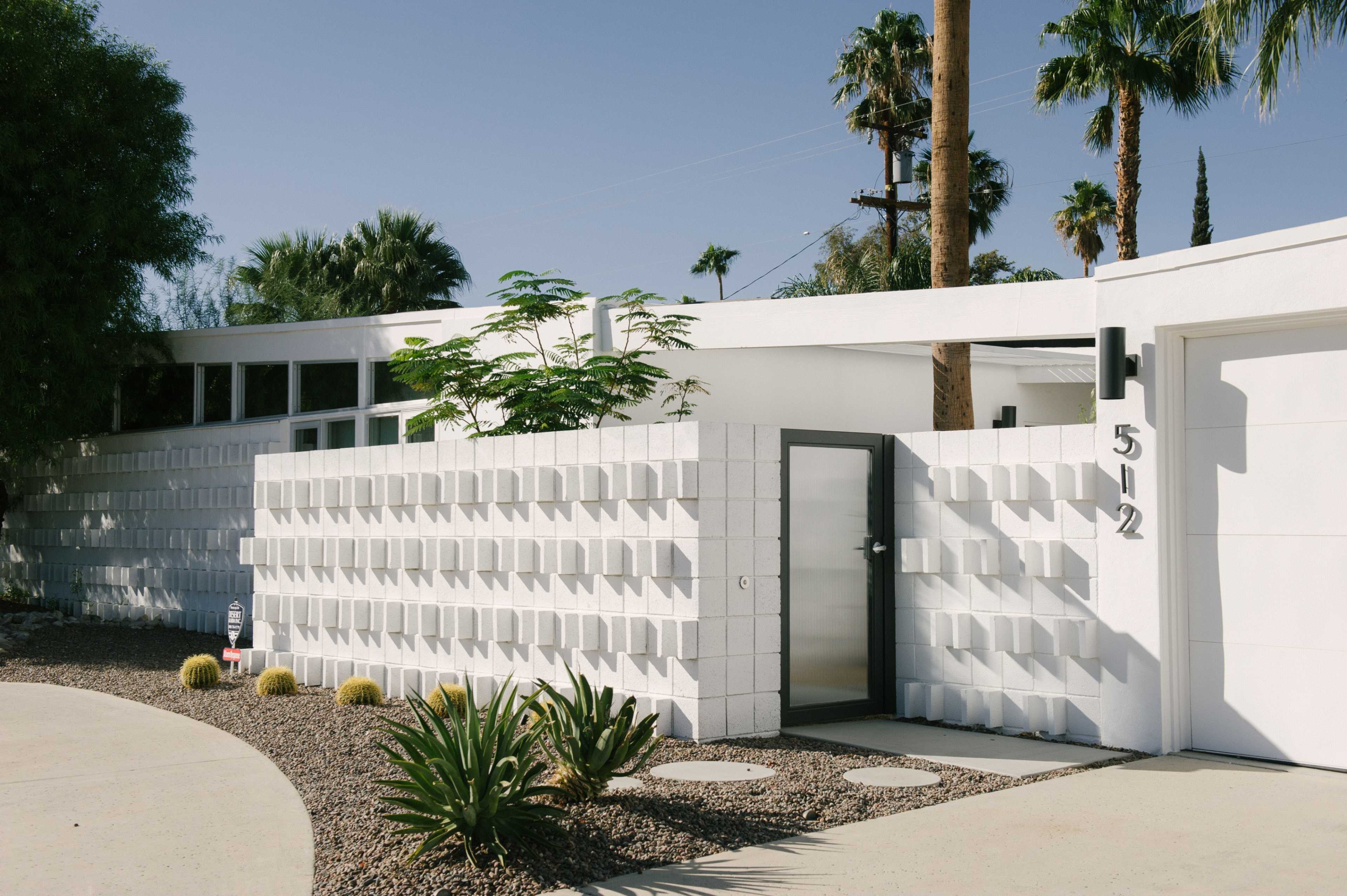 The image shows a modern white house with a textured concrete wall and a glass door, surrounded by desert landscaping and palm trees.