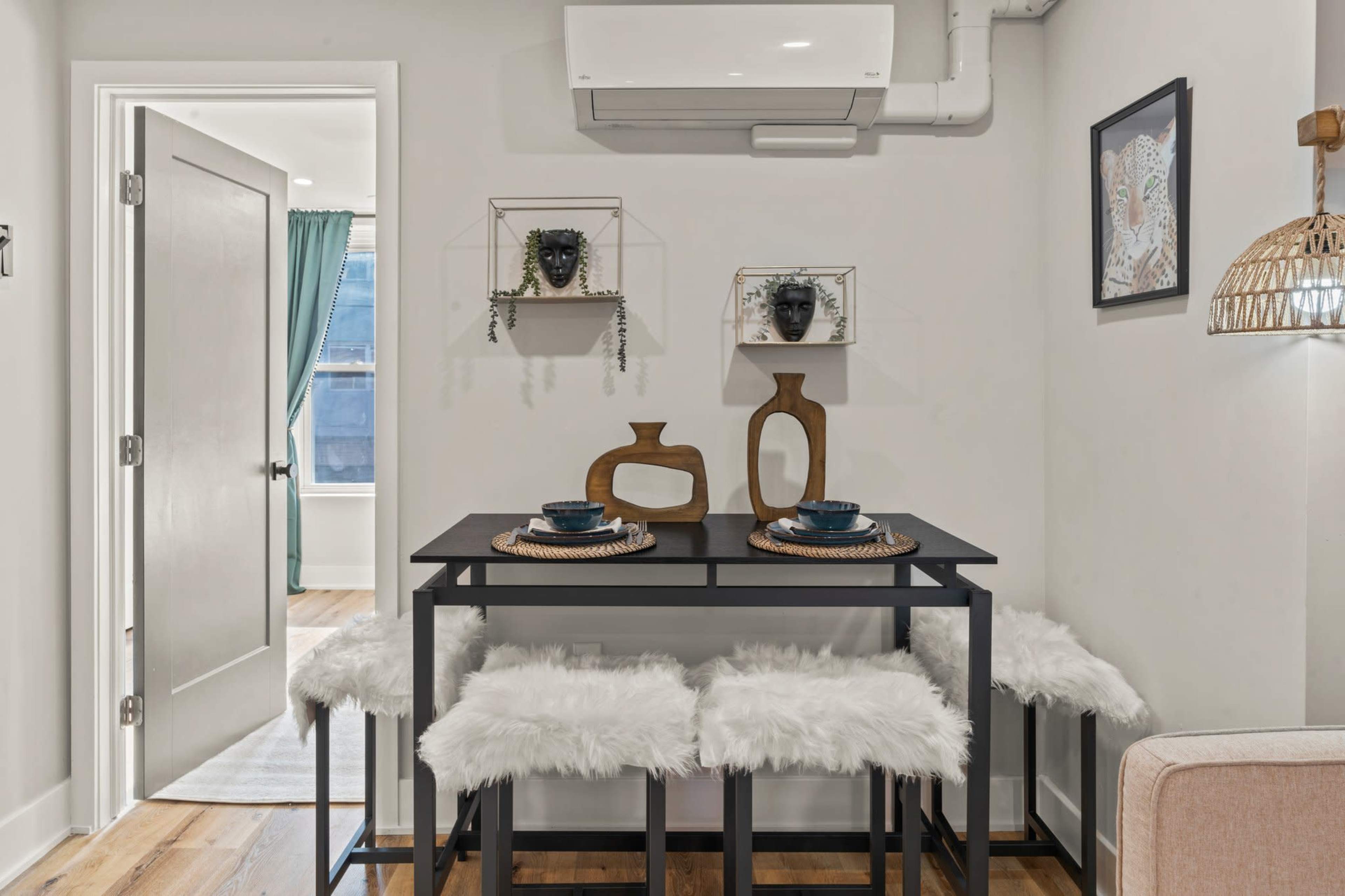 A small dining area features a black table with two wooden decorative pieces and chairs covered in white fur, alongside a door leading to another room.