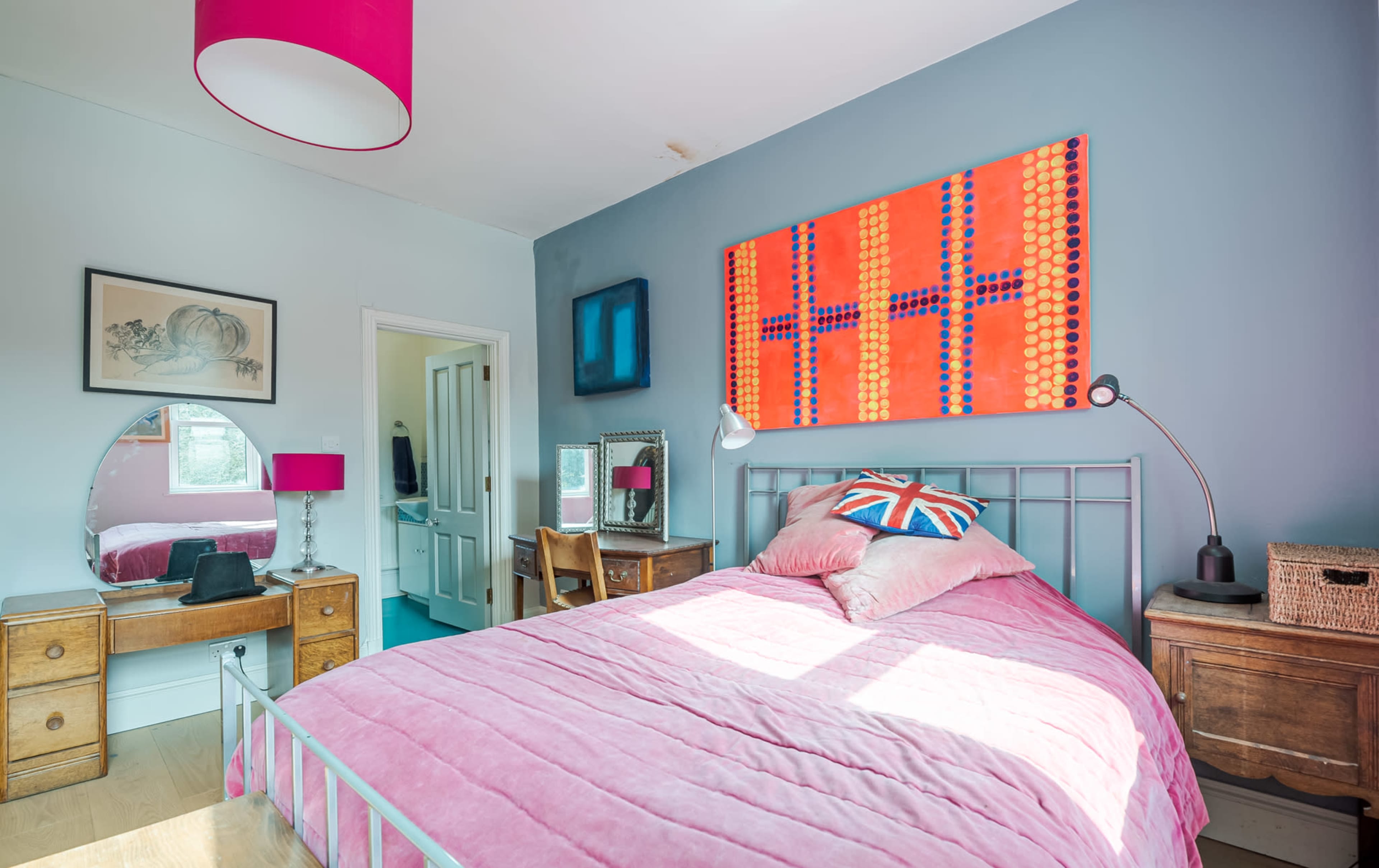 The room features a pink bedspread on a metal bed frame, with a colorful artwork above it, a round mirror, and wooden furniture, including a chair and a basket.