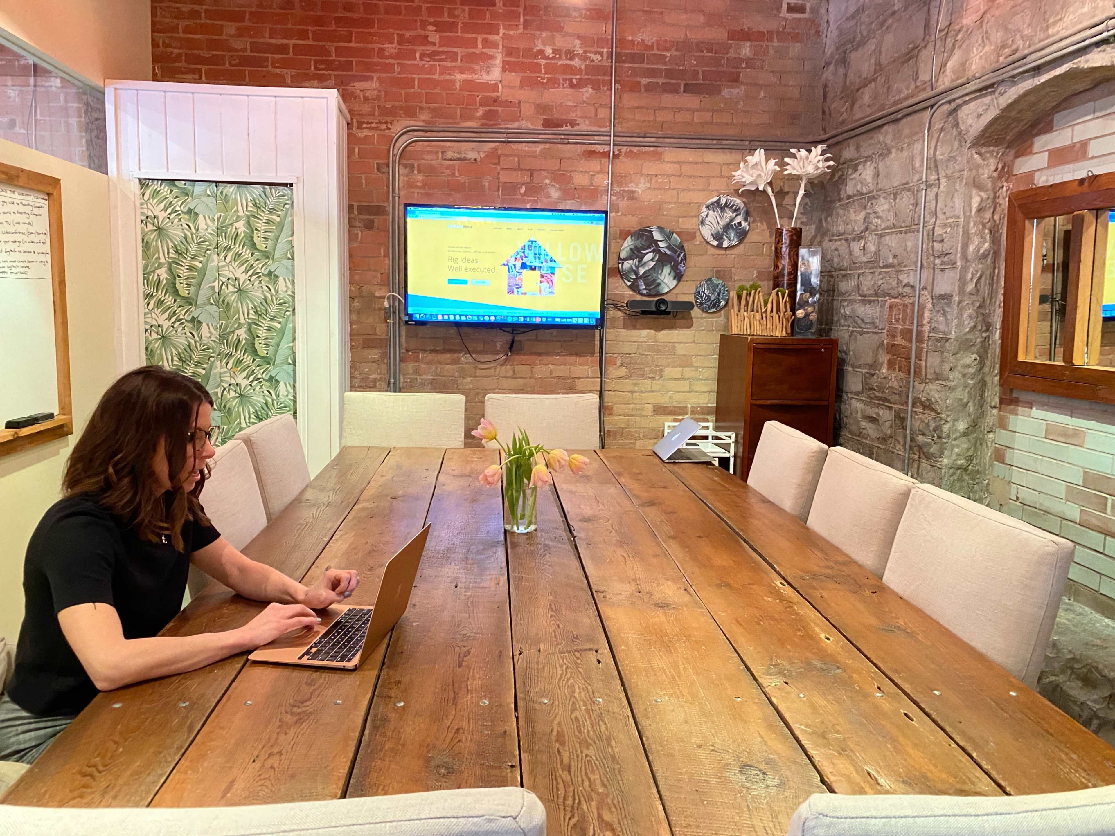 A person is working on a laptop at a long wooden table in a meeting room with a large screen displaying a colorful website in the background.