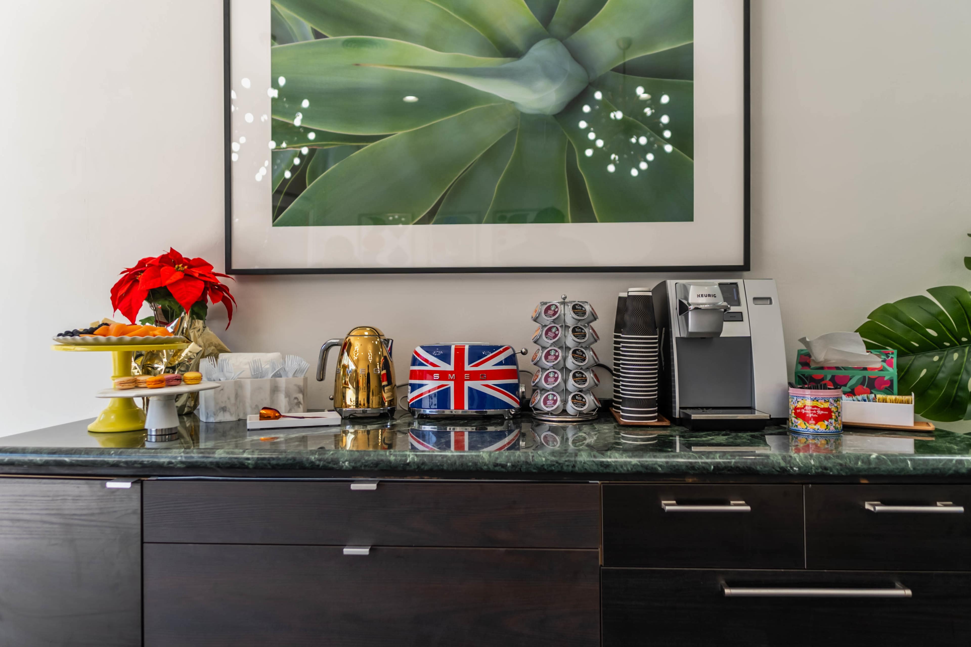 A countertop features a coffee machine, cups, a teapot, snacks on a tiered tray, and decorations including a British flag and a poinsettia, all against a backdrop of a large plant print.