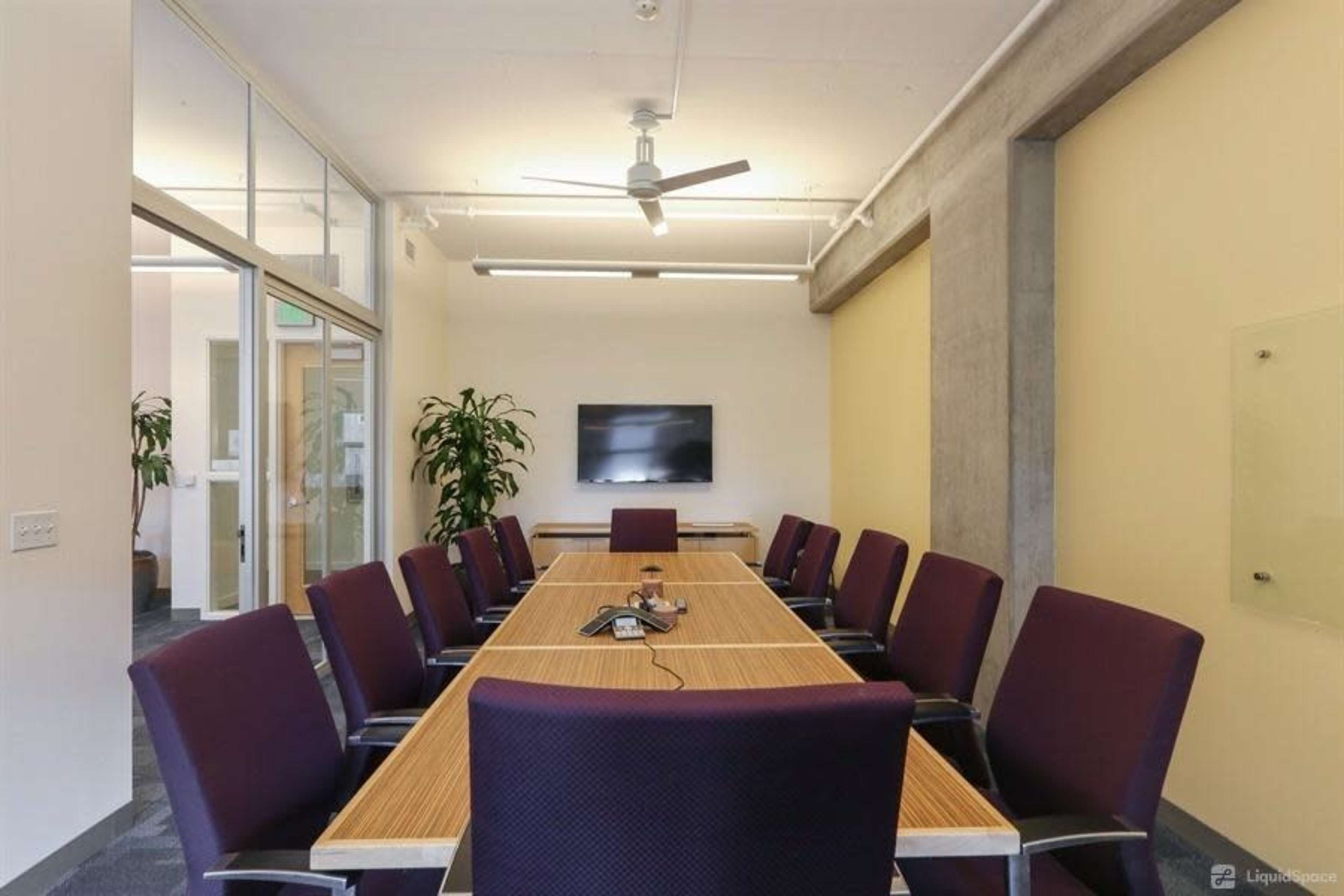 A conference room features a long wooden table surrounded by purple upholstered chairs, with a television mounted on the wall and plants in the corners.