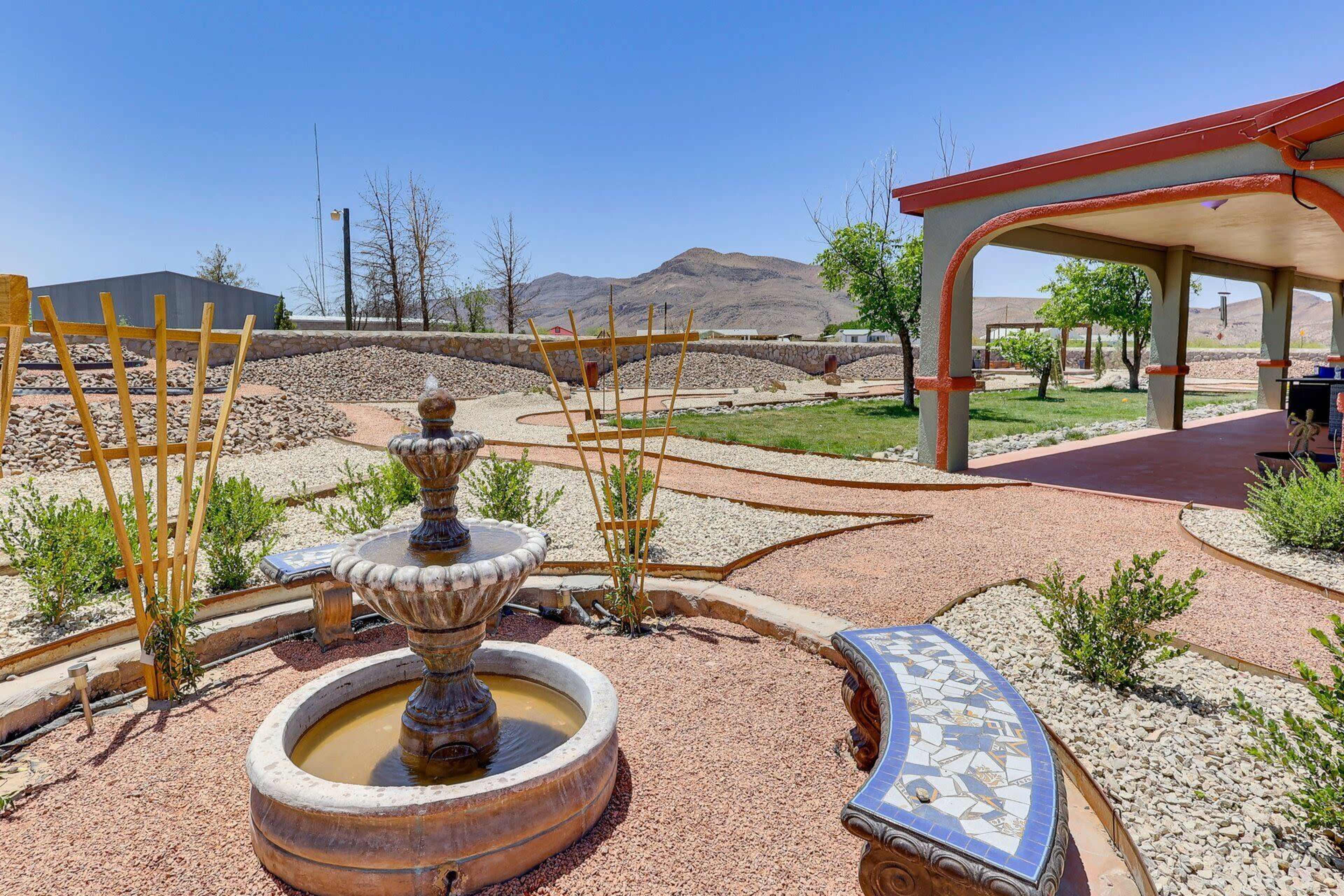 The image features a decorative fountain surrounded by landscaped pathways and shrubbery, with a mountain view in the background.