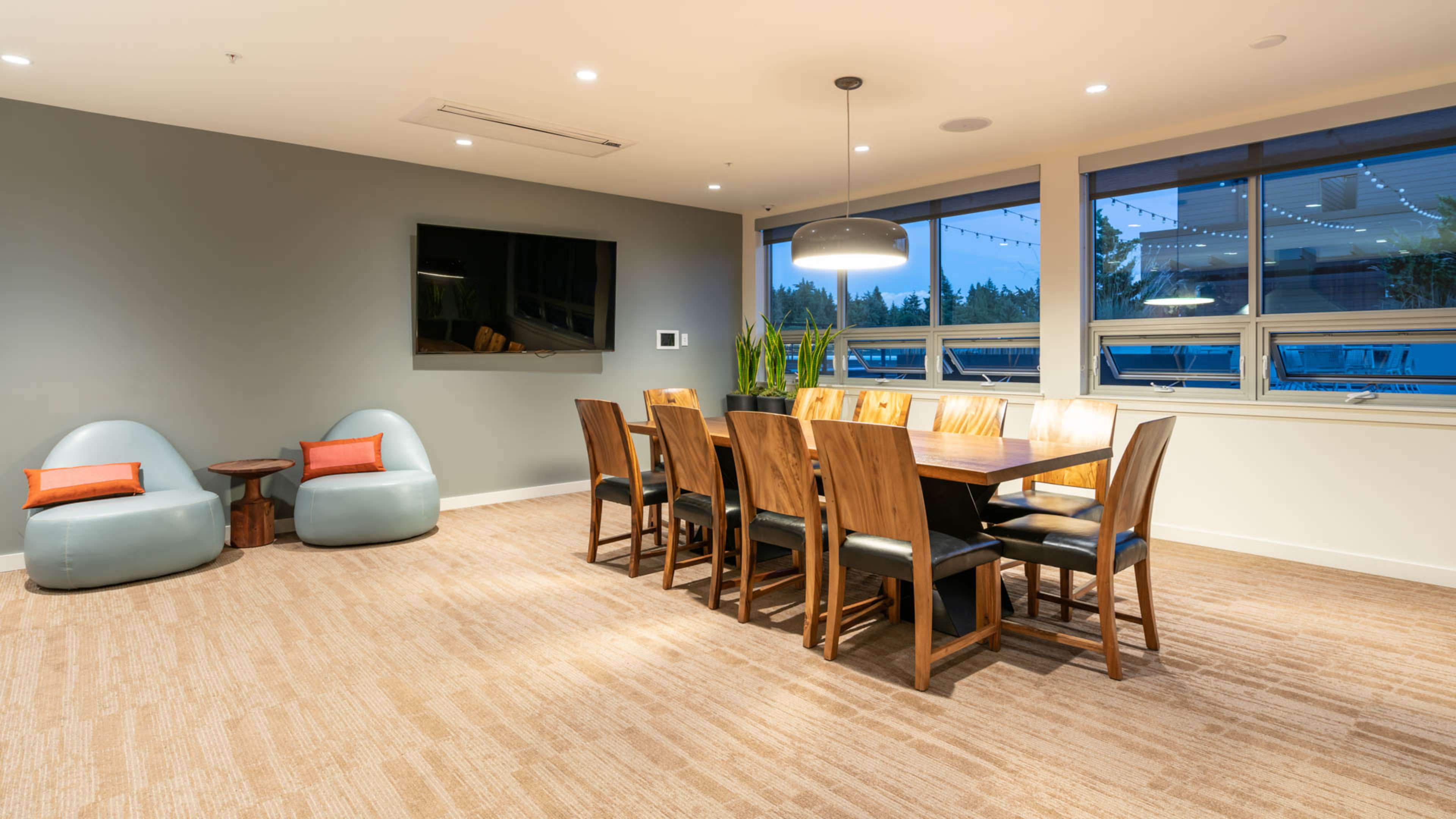 A modern dining area featuring a long wooden table surrounded by chairs, a large television on the wall, and two cushioned seats in a corner.