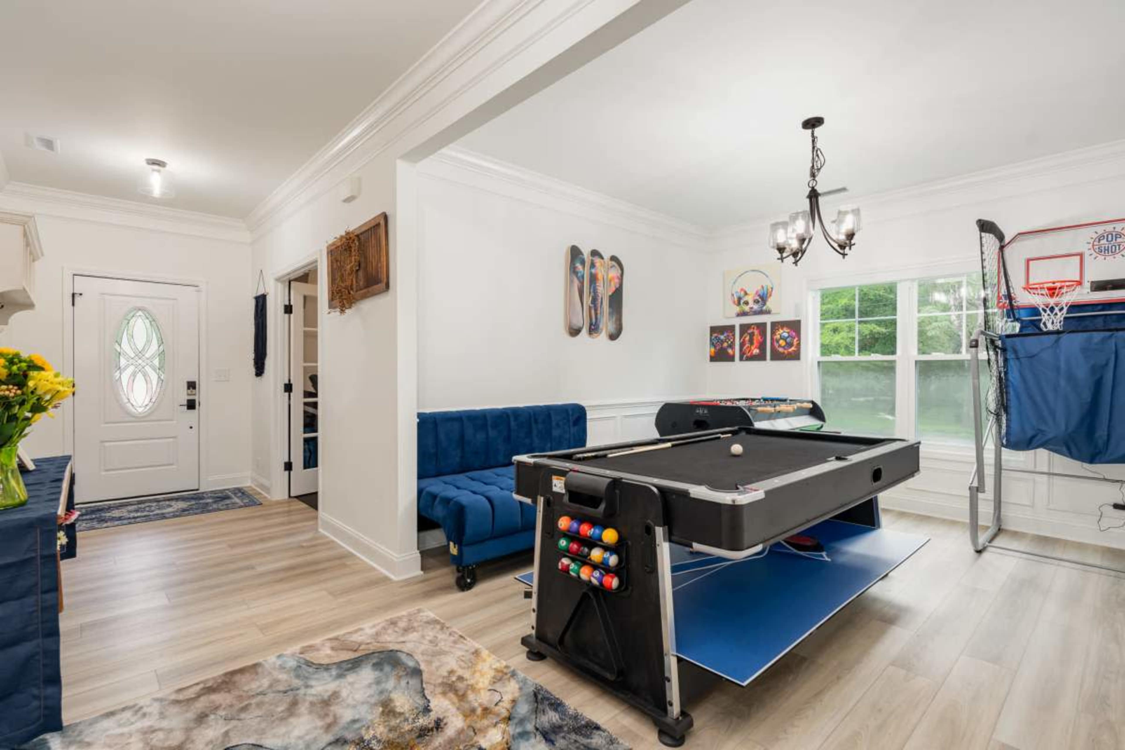 The room features a pool table, a blue couch, and a basketball hoop, with a door and large windows providing natural light.
