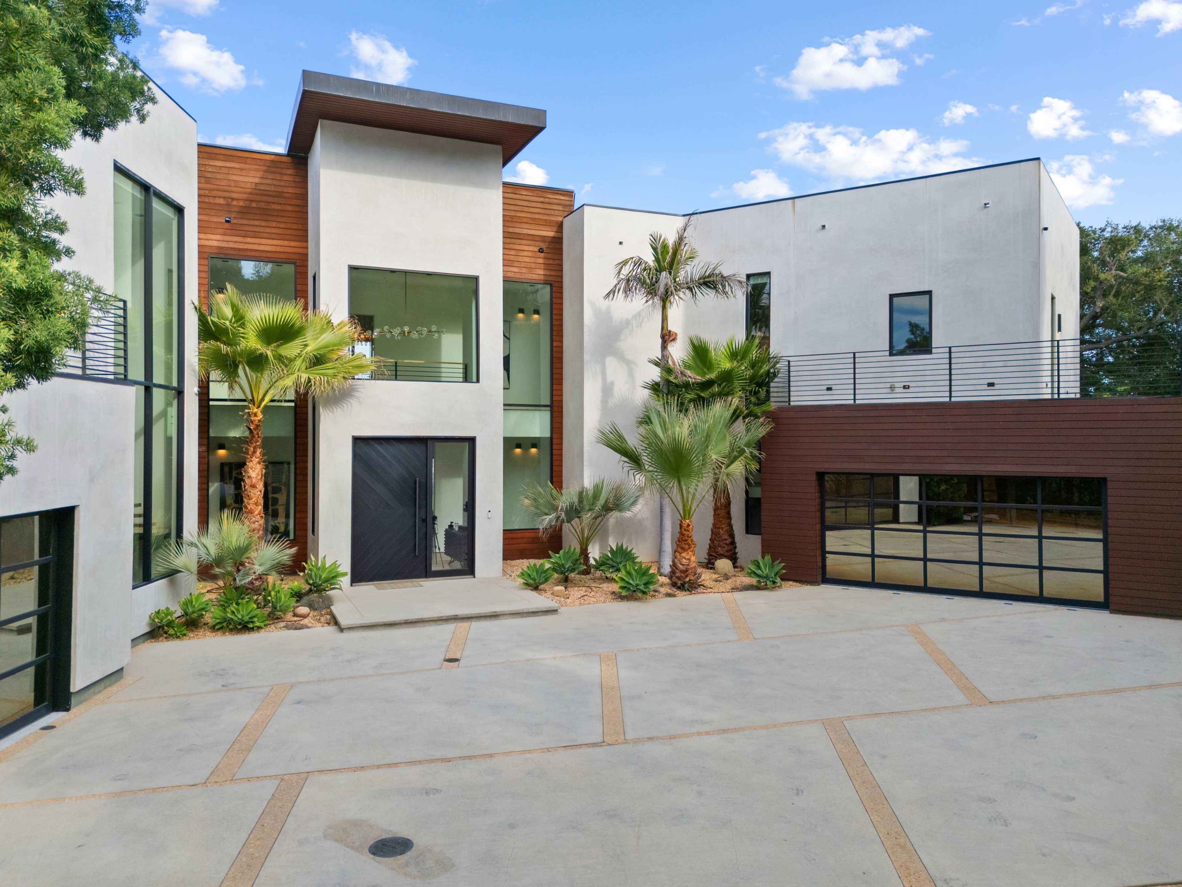 The image shows a modern two-story house with large windows, a mix of wood and stucco exteriors, palm trees, and a spacious driveway.