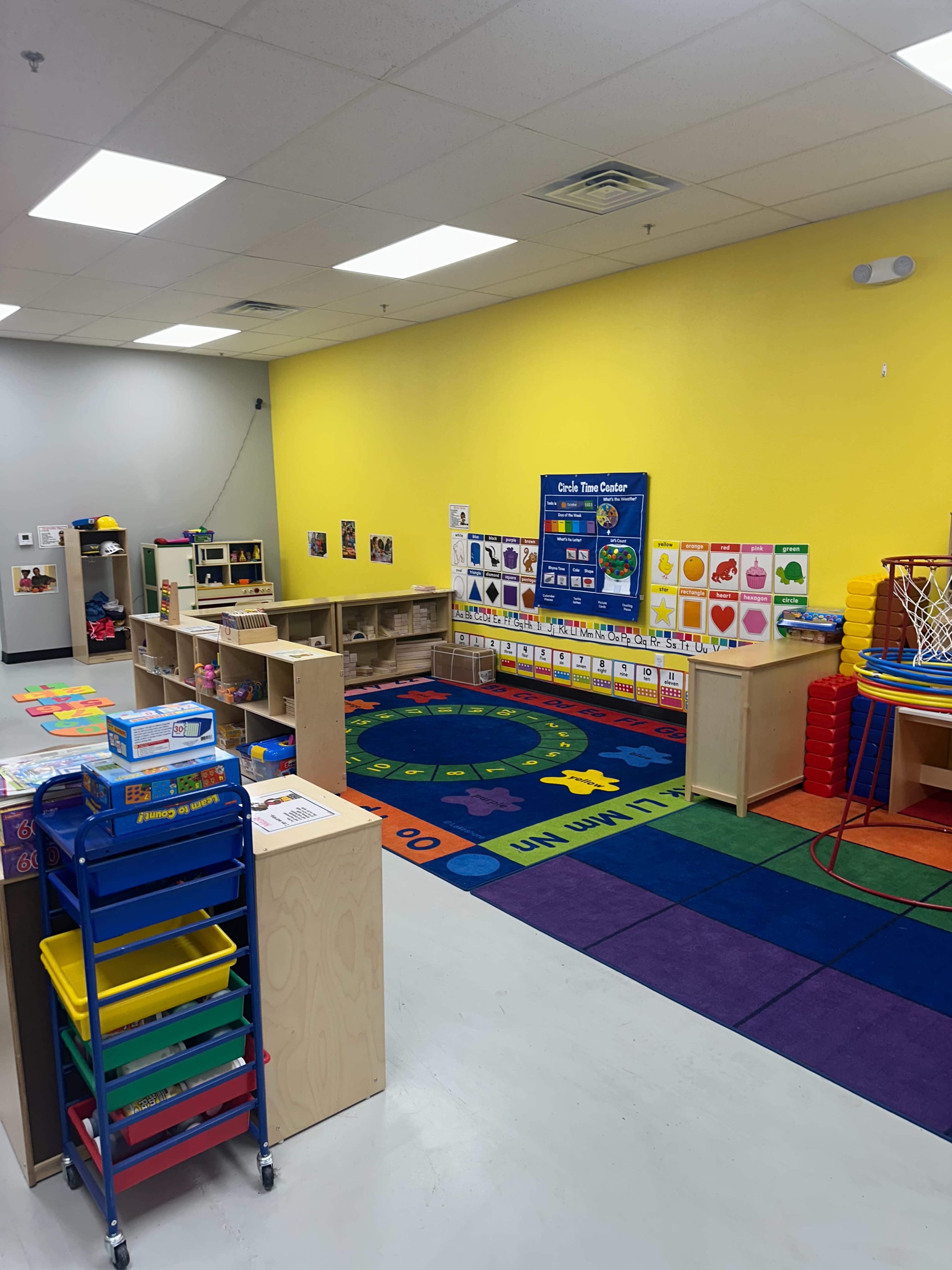 The image shows a brightly colored classroom with various learning materials, including shelving units, rugs, and a bulletin board on a yellow wall.