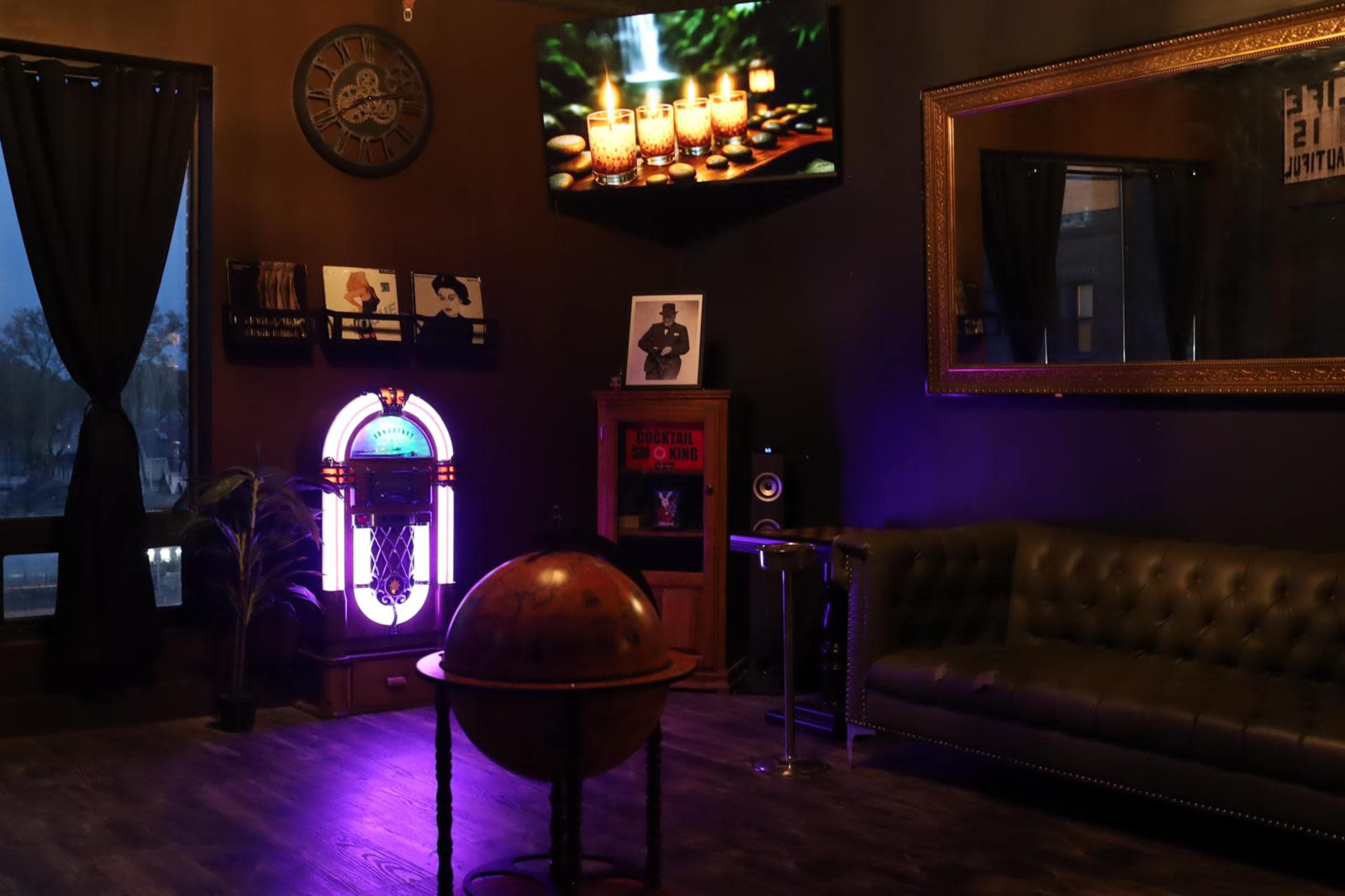 The image shows a dimly lit lounge area featuring a globe in the center, a jukebox, a sofa, and a television displaying candles in a serene setting.