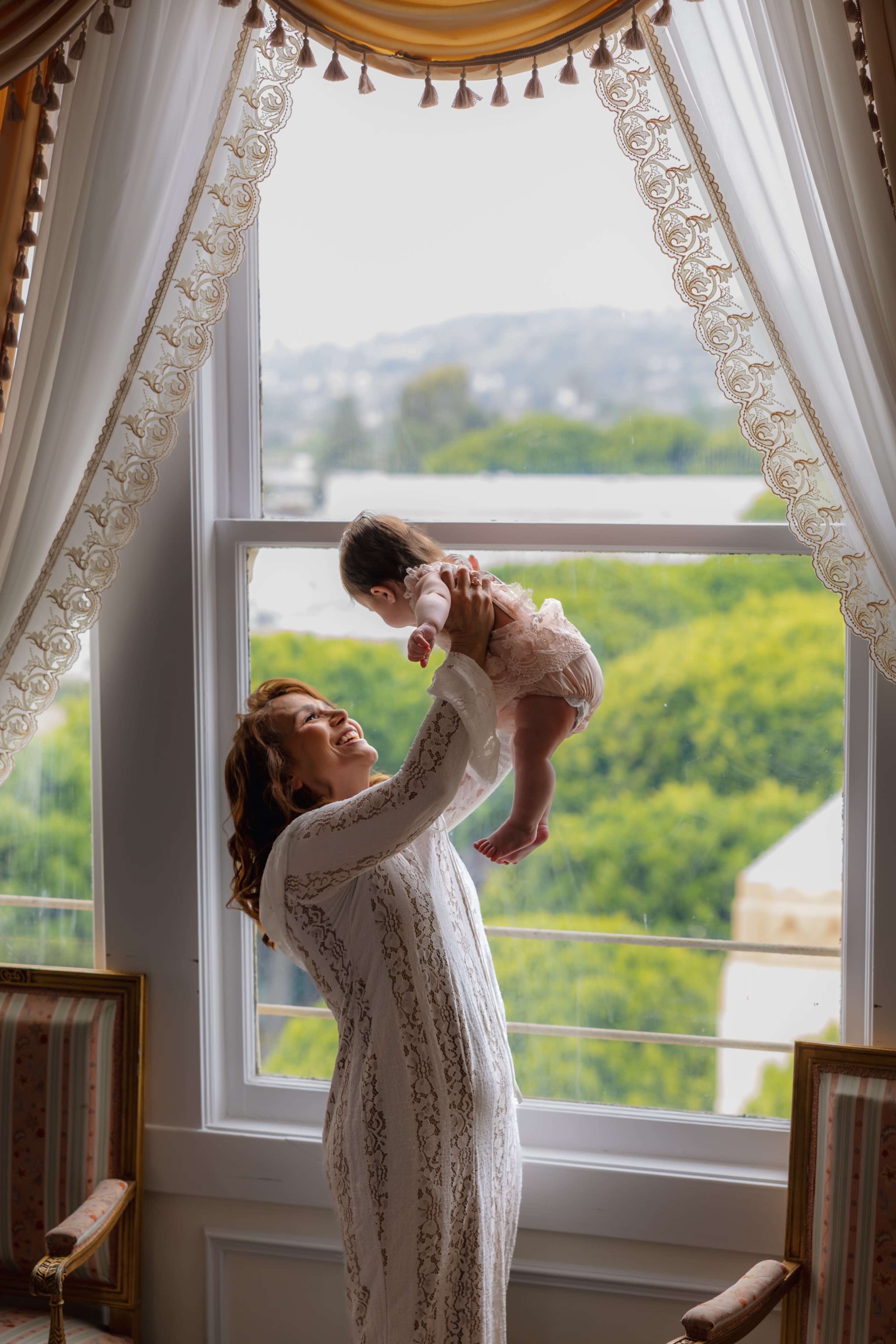 A woman in a white dress lifts a baby toward a large window with a view of greenery outside.