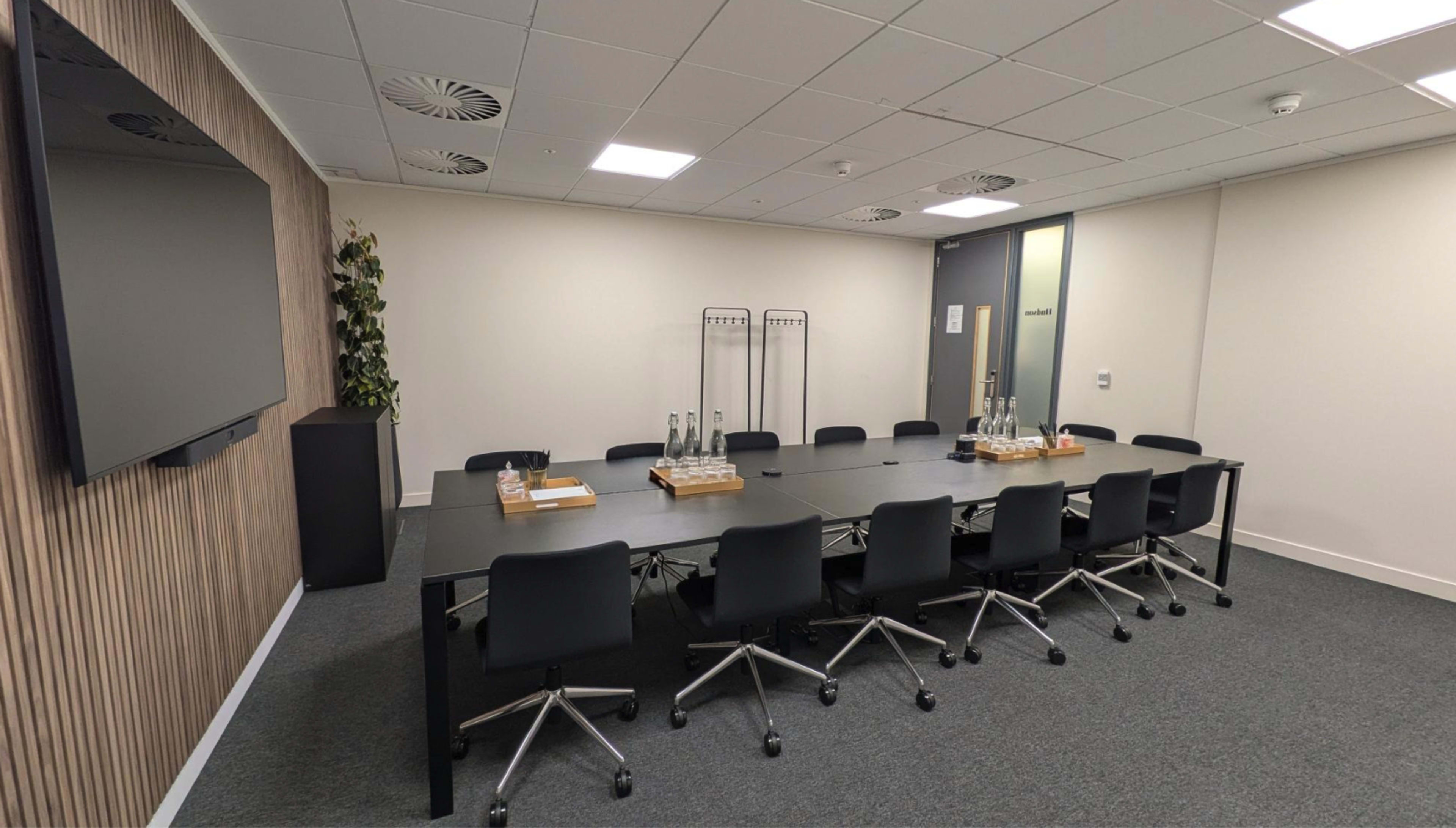 A modern conference room features a large black table surrounded by rolling chairs, with a television mounted on the wall and water bottles on the table.