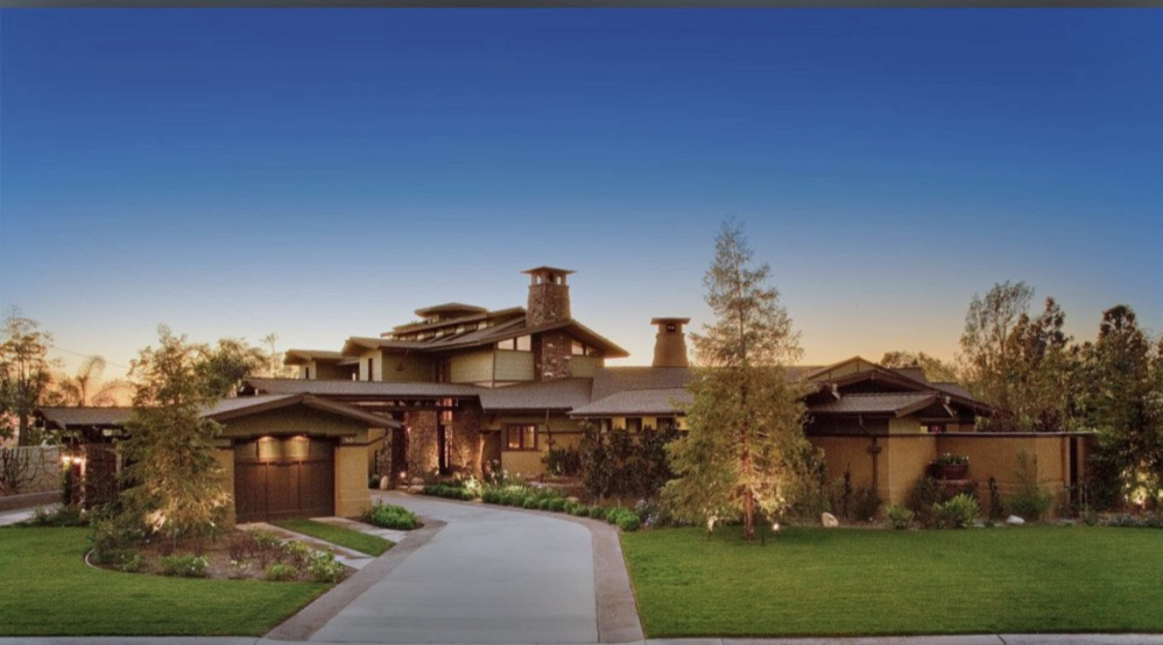 A modern two-story house with a stone chimney and a curved driveway, surrounded by landscaped greenery and trees, under a clear blue sky.