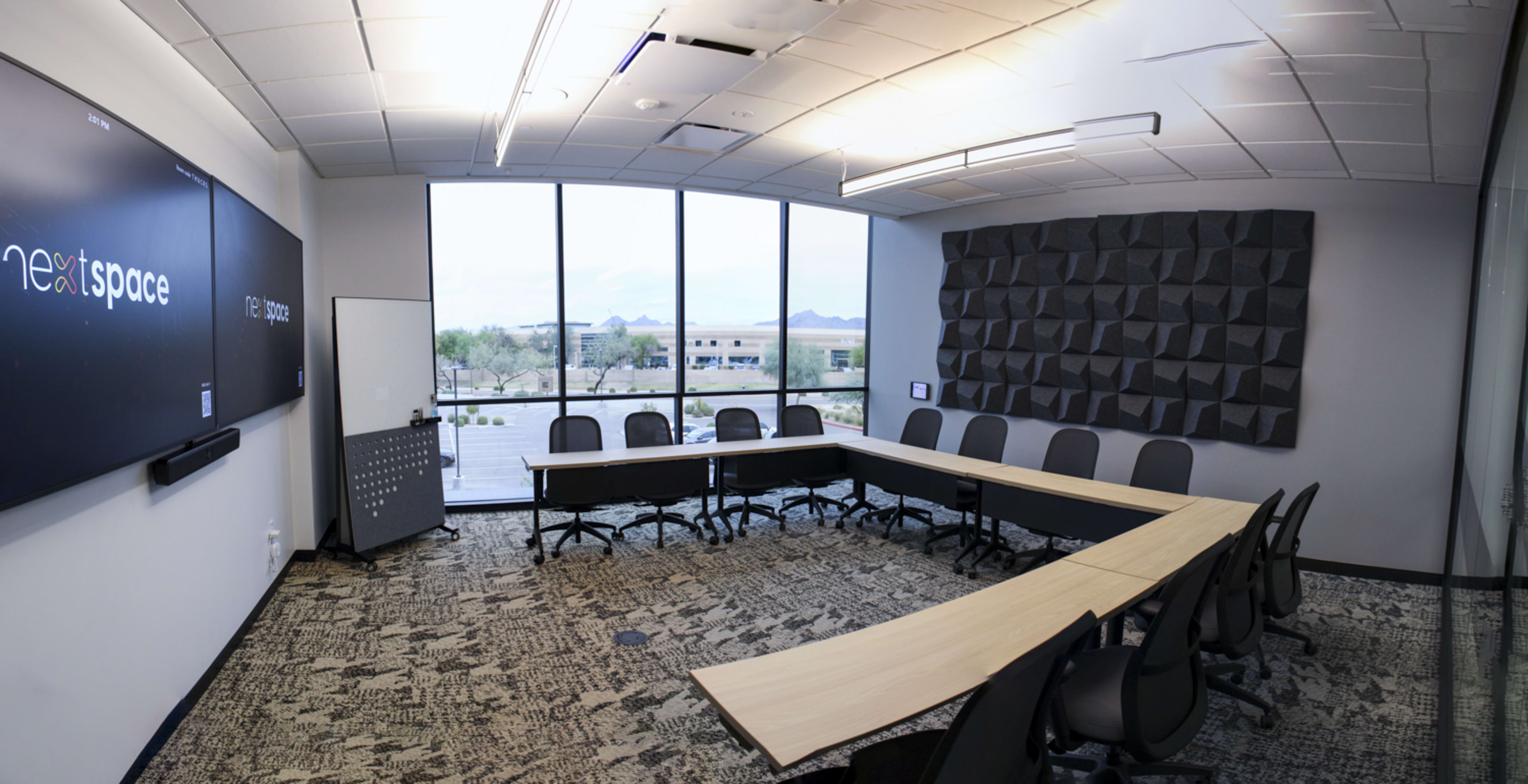 The image shows a modern conference room with a long table, black ergonomic chairs, a large screen, and windows displaying a view of the landscape outside.