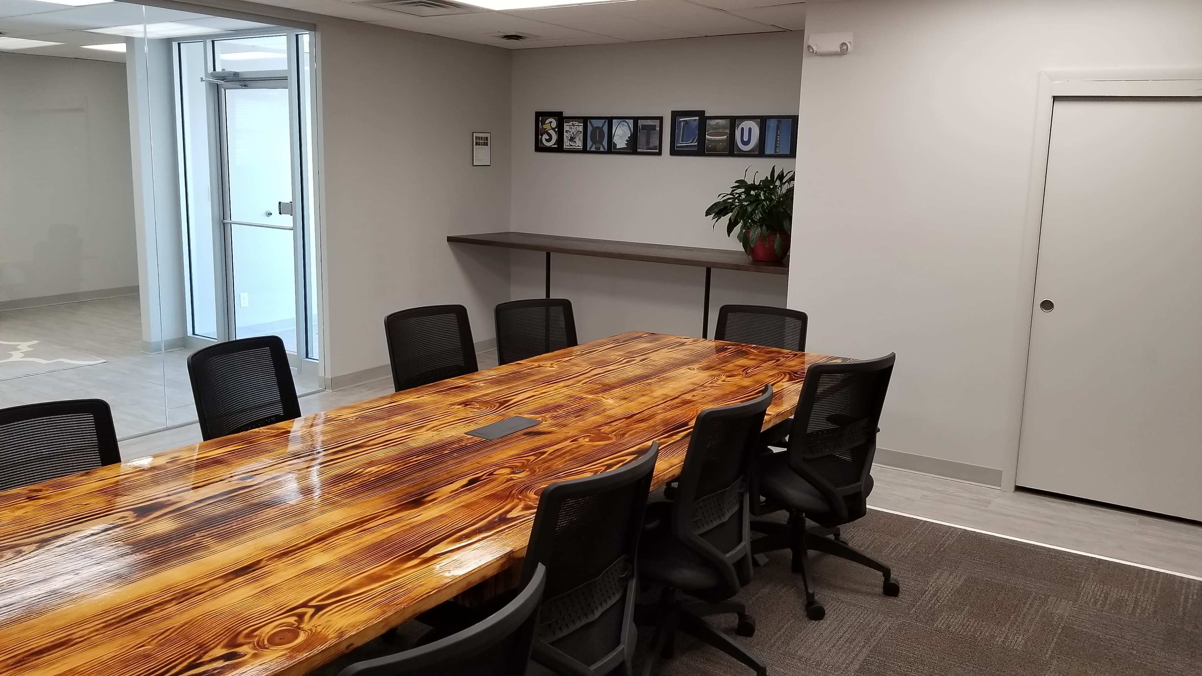 A large wooden conference table is centered in a meeting room with black chairs, while a wall display featuring framed awards and a potted plant is visible in the background.