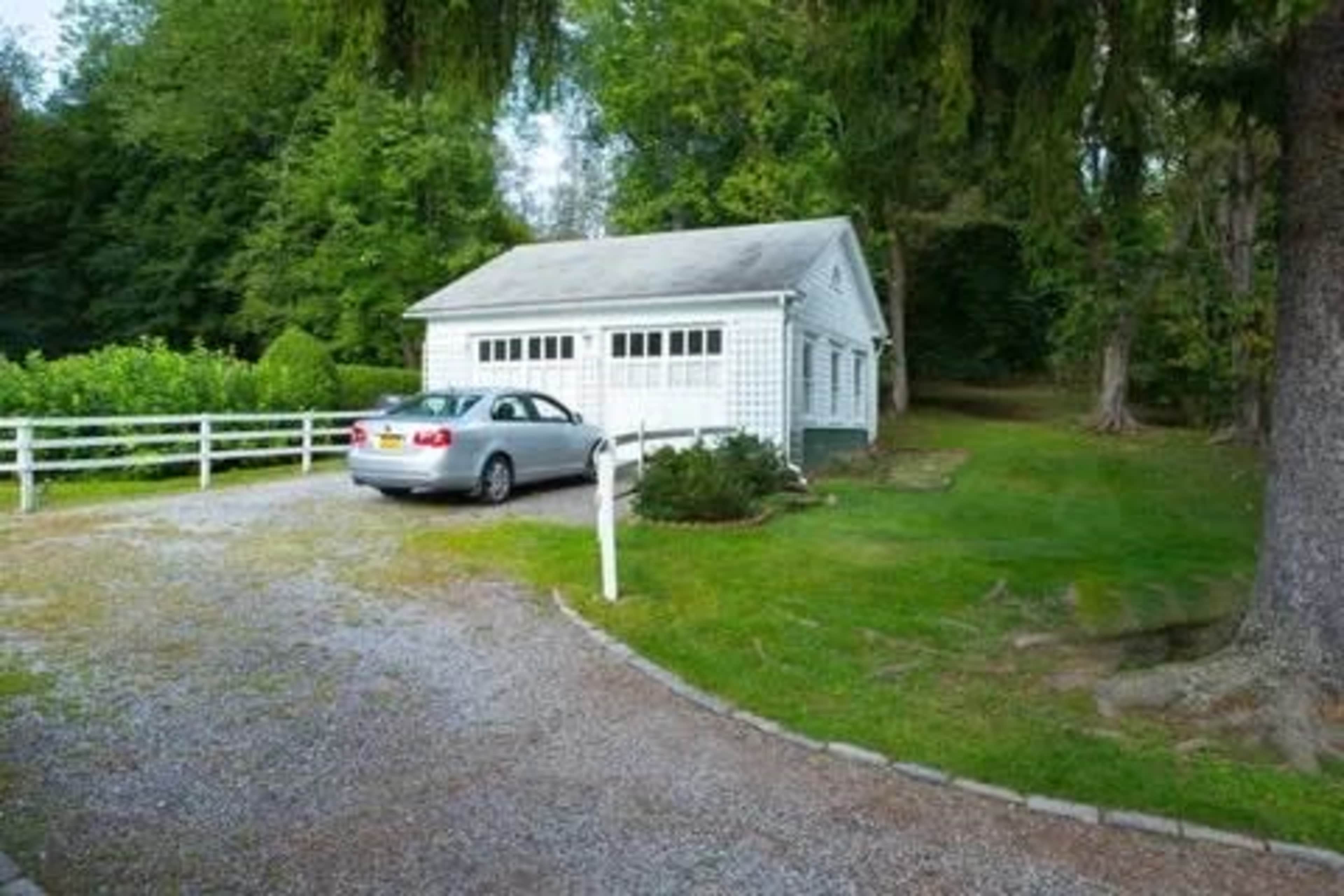 A gray car is parked beside a white garage on a gravel driveway surrounded by greenery.