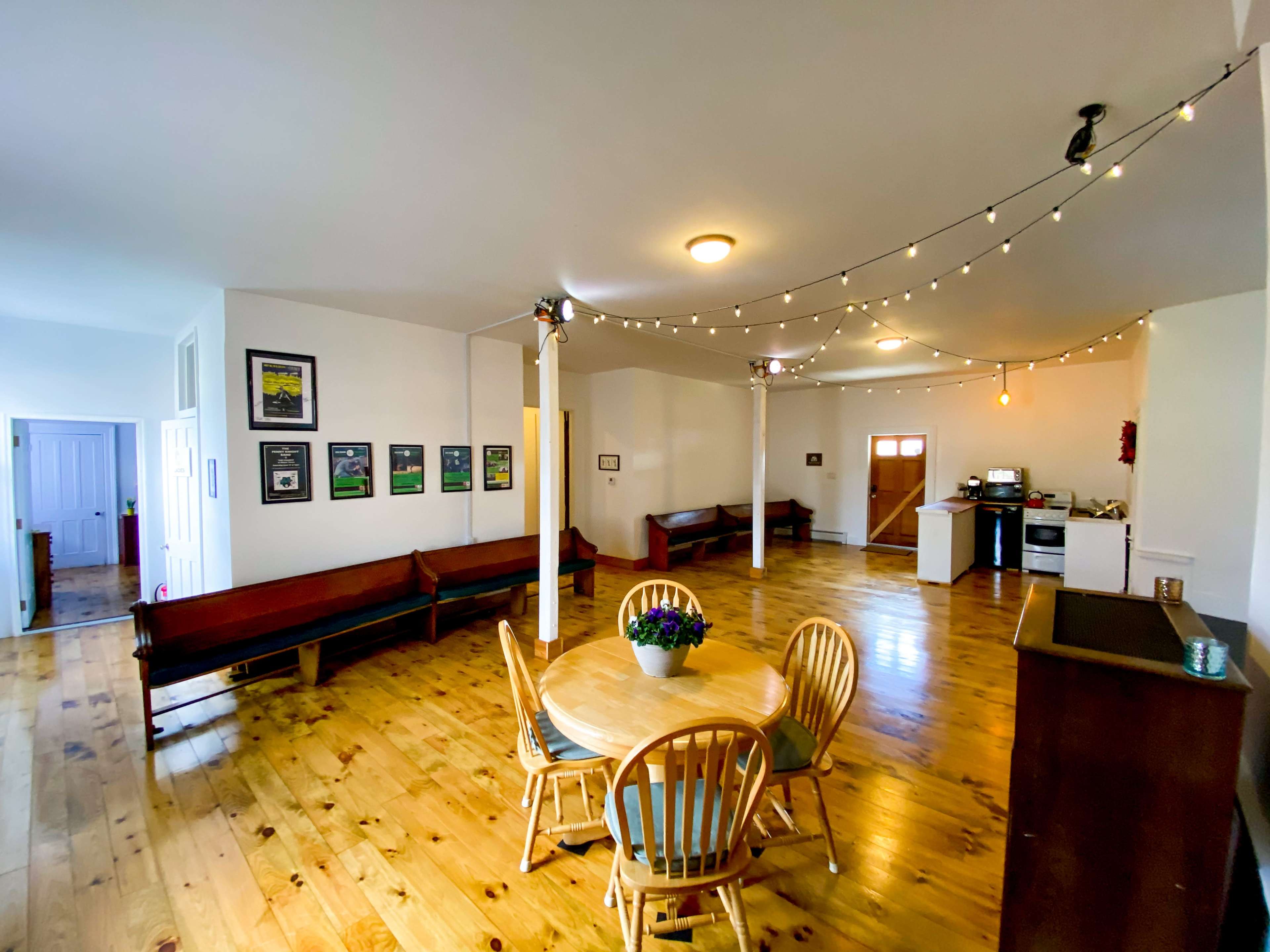 The image shows a spacious, well-lit interior featuring a dining area with a round table and chairs, wooden floors, and a kitchenette in the background.