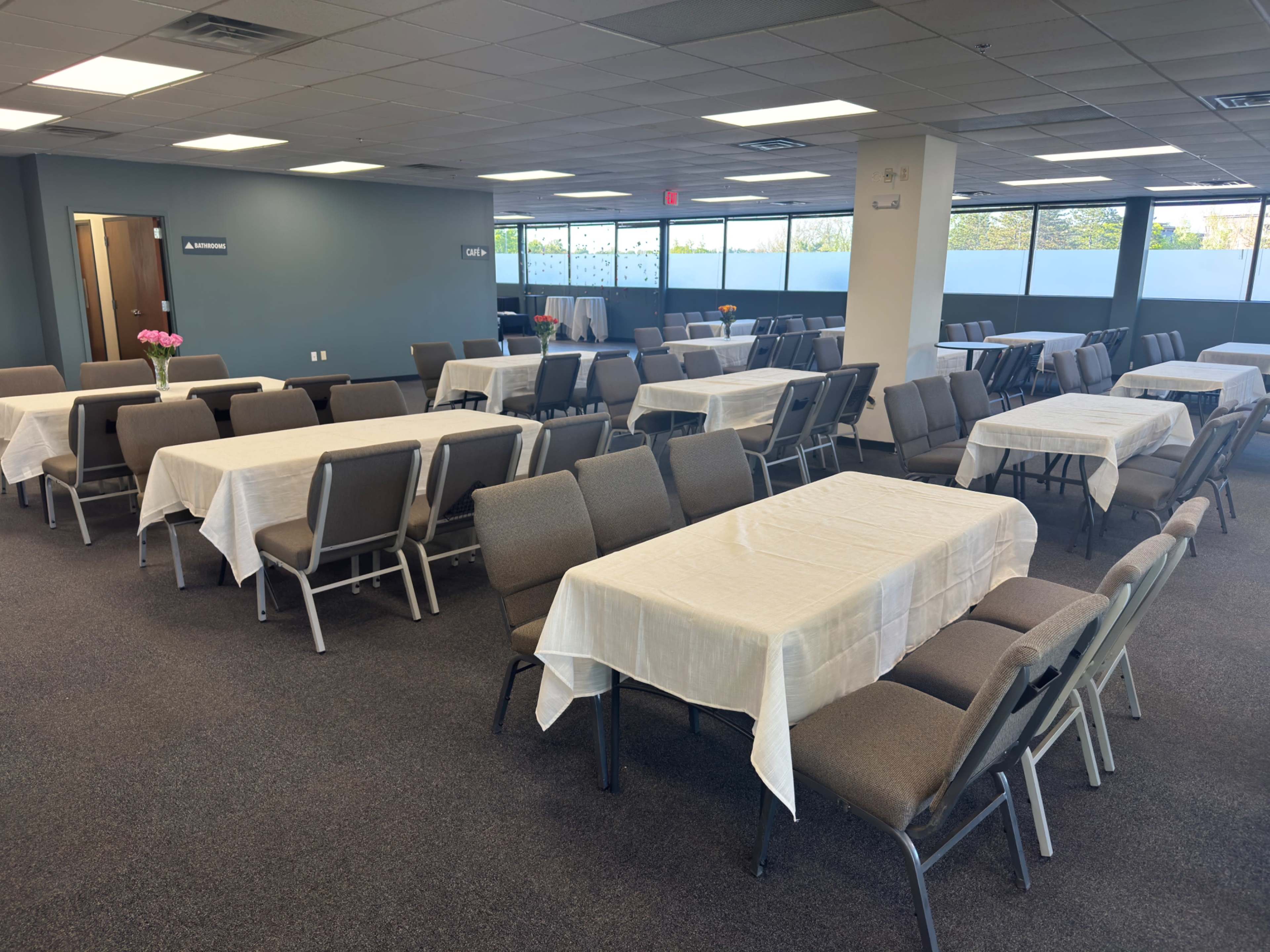 The image shows a spacious room arranged with multiple tables covered in white tablecloths and surrounded by chairs, ready for an event.