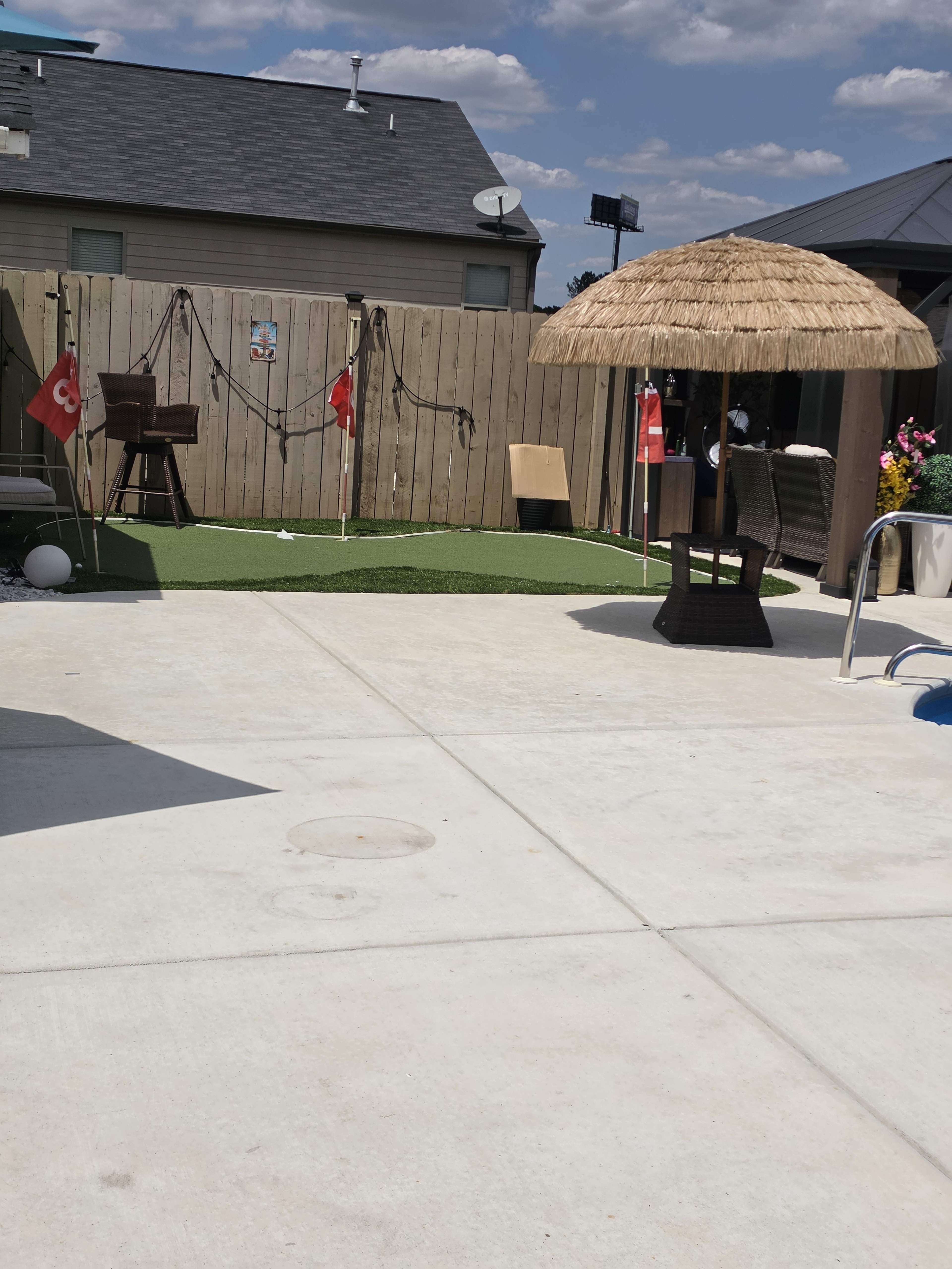 The image shows a pool area with concrete flooring, a thatched-roof gazebo, artificial grass, and several decorative flags along a wooden fence.