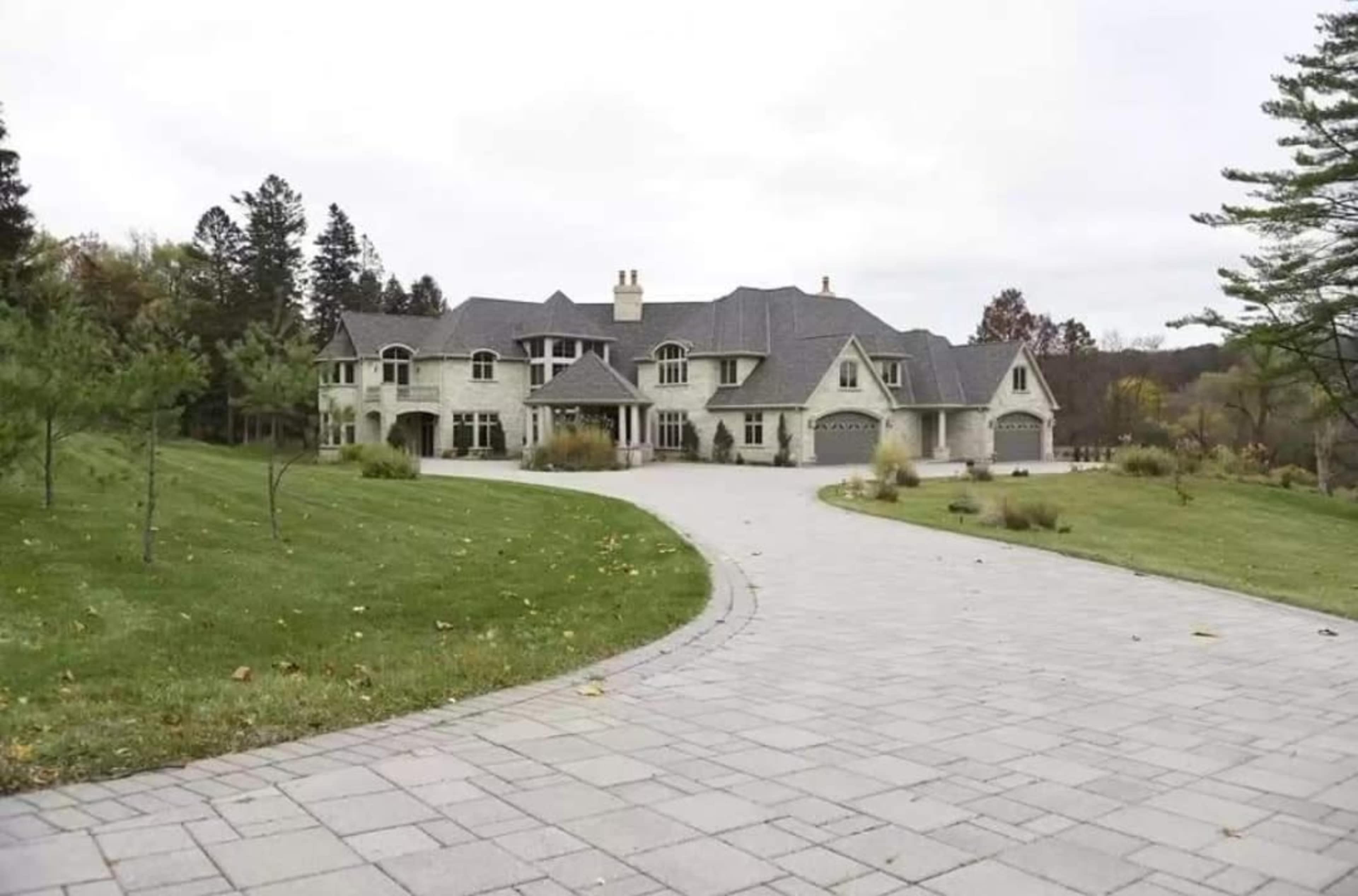 A large two-story mansion with a stone exterior and a paved driveway is surrounded by manicured lawns and trees.