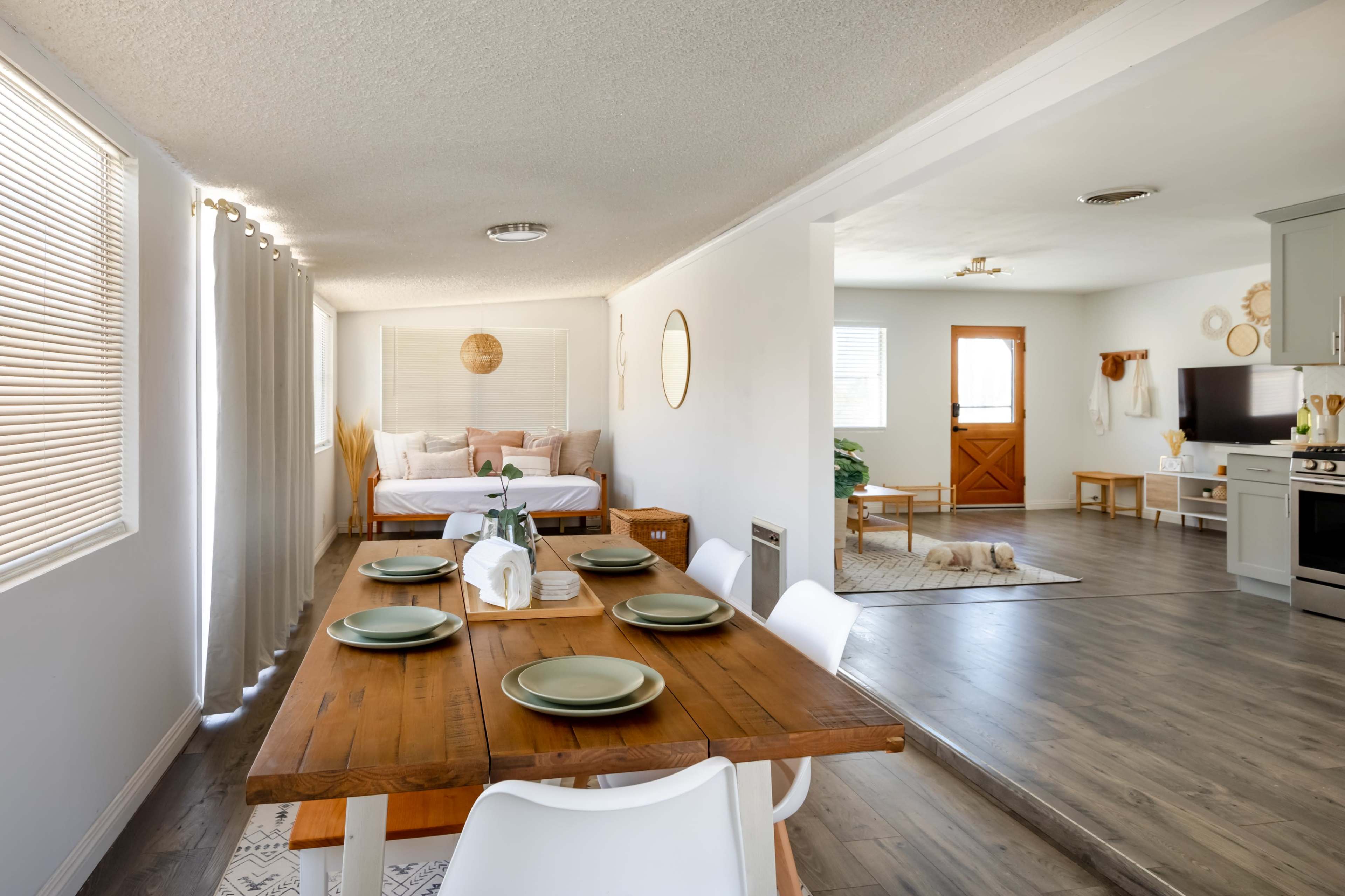 The image shows a modern, open-concept living space with a dining area featuring a wooden table set for four, leading to a small bedroom area with a bed and a living area with a television.
