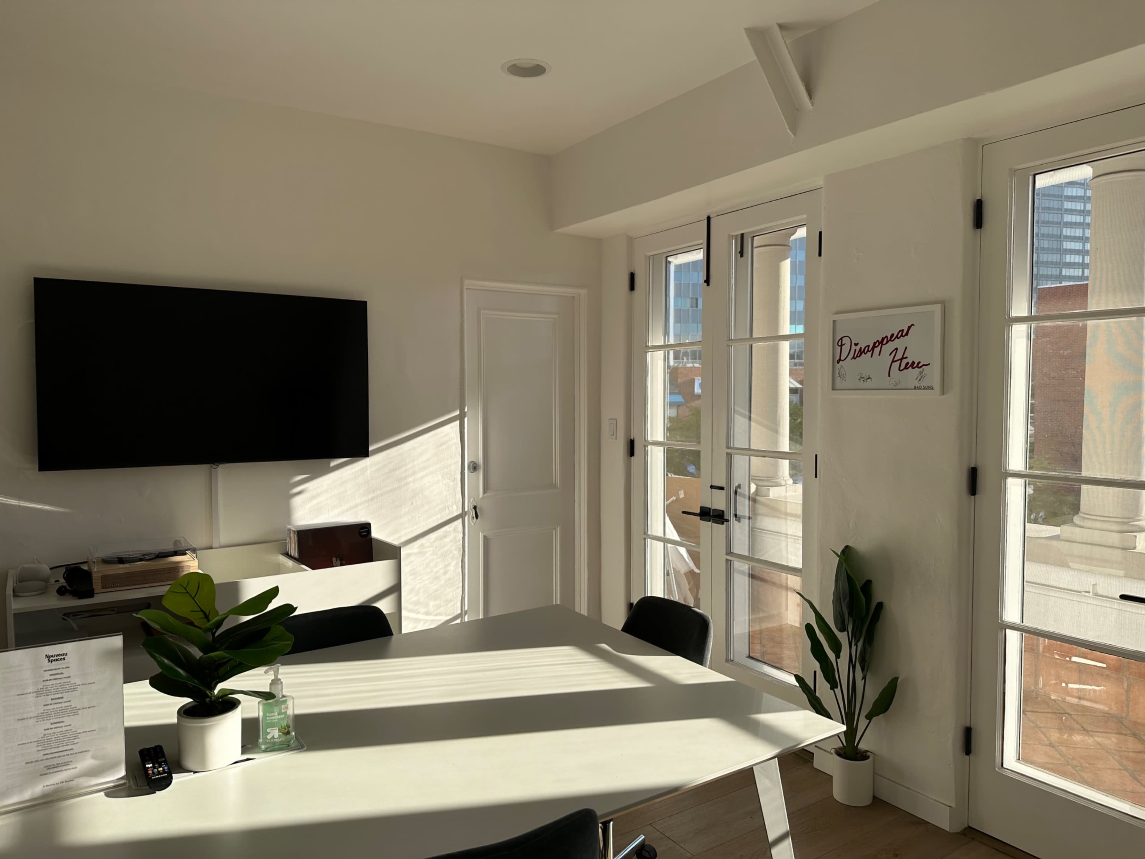 A well-lit office features a modern table, a TV mounted on the wall, and large windows with plants by the side.
