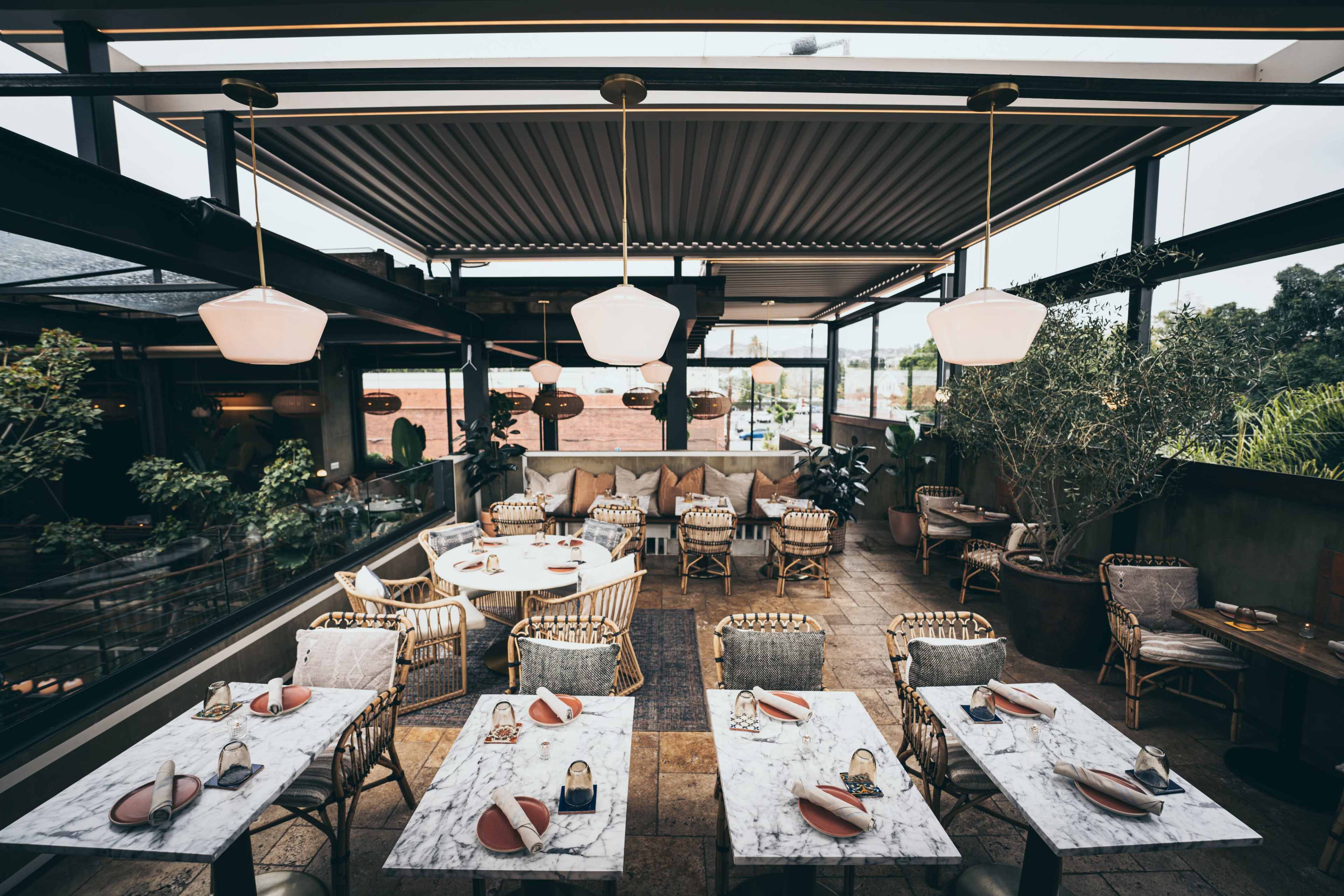 A modern restaurant interior with several tables set for dining, surrounded by greenery and large windows.