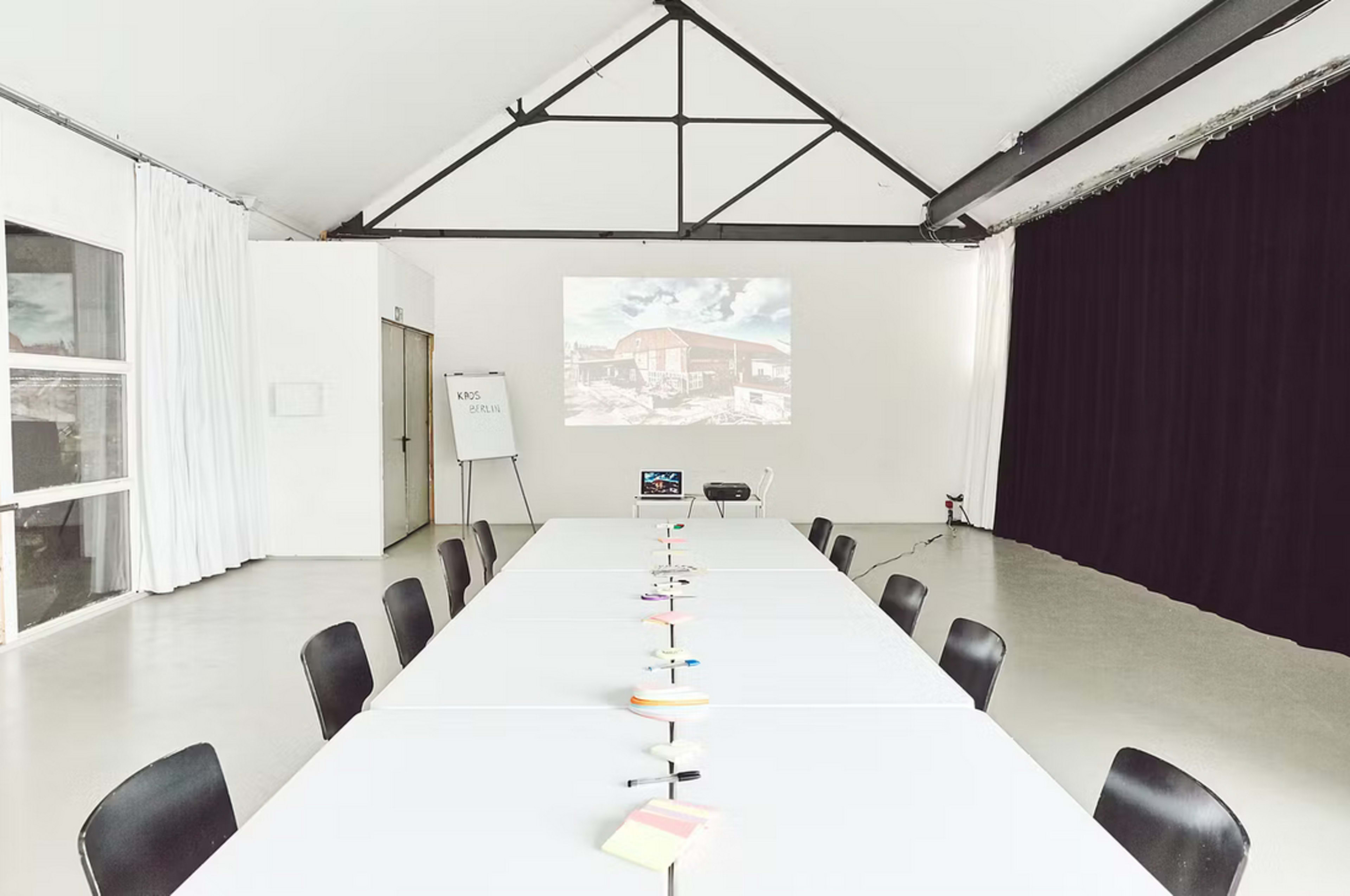 A long, white conference table is set up in a meeting room with black chairs, and a projector displays an image on the wall.
