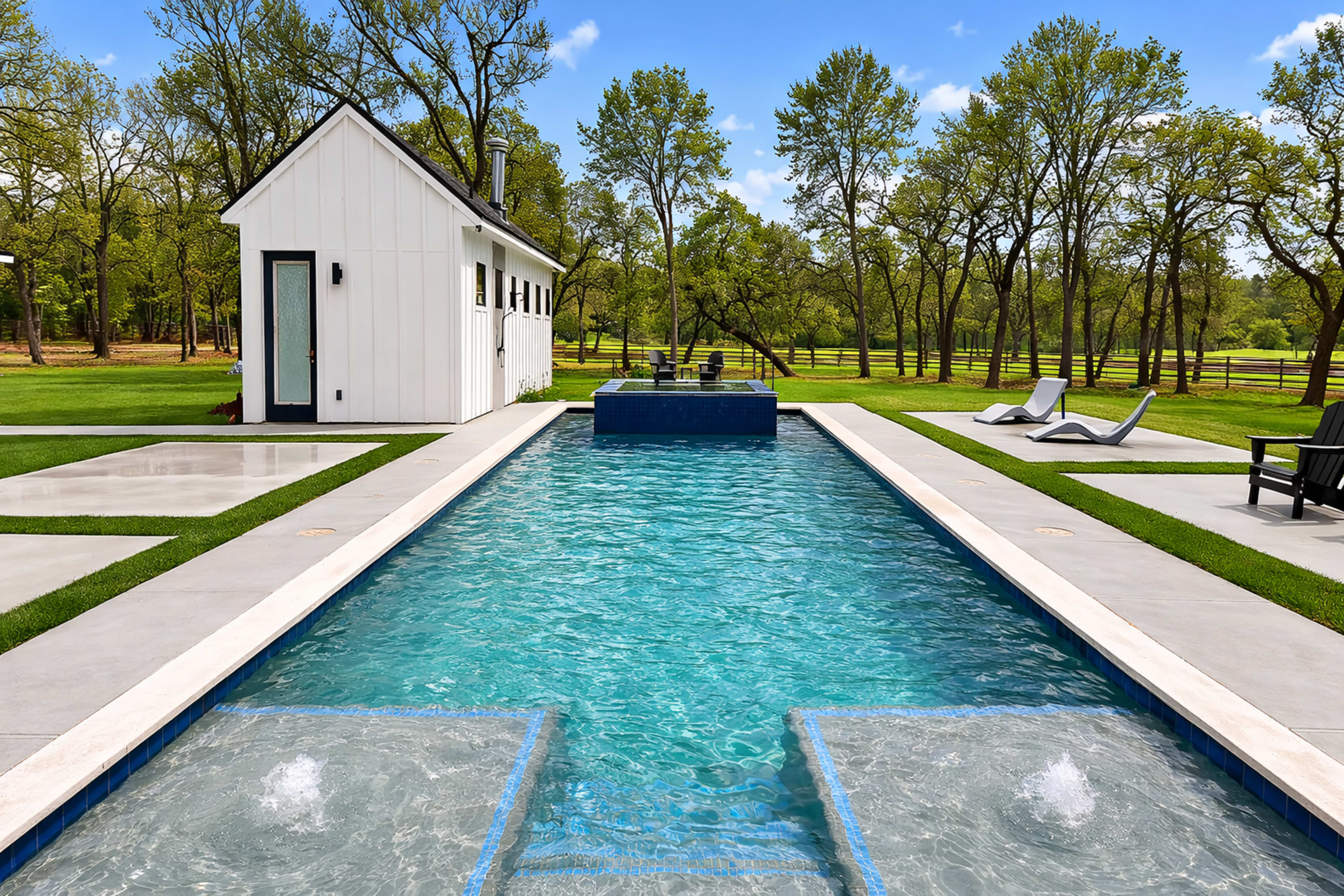 A rectangular swimming pool is lined with concrete, surrounded by green grass and white lounge chairs, with a small white shed in the background.